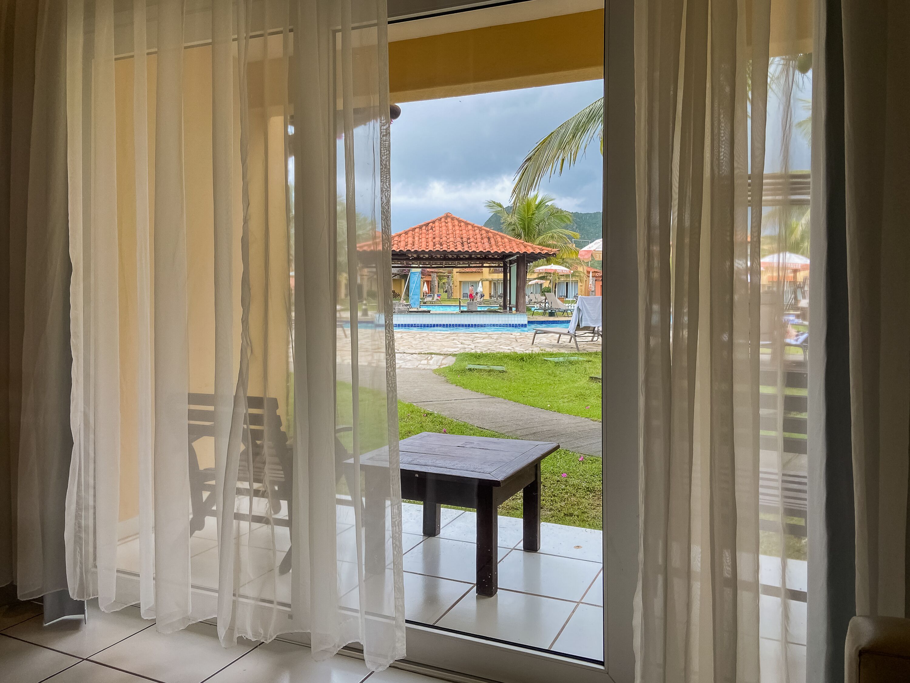 A room with a view of a pool area, including a covered seating area and a small table.