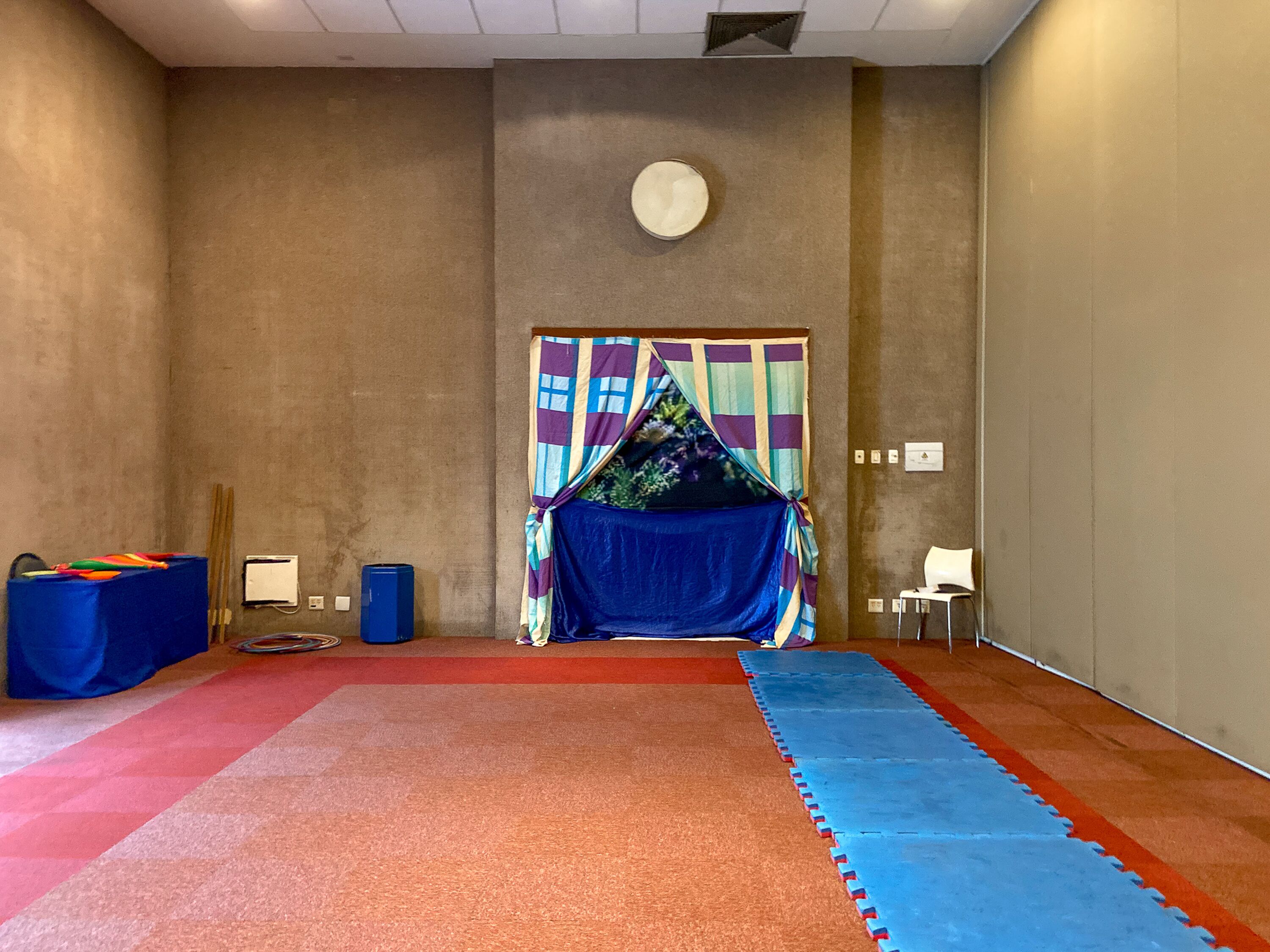 Una habitación con alfombra roja, tapete azul, mueble de almacenamiento azul y cortina colorida.