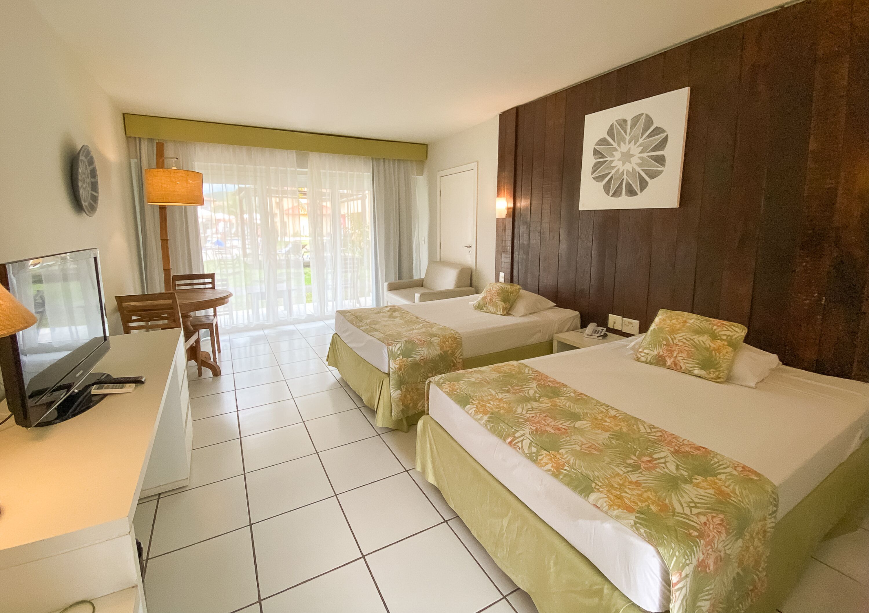 A hotel room with two beds, a desk with a television, a chair, and a large window with curtains.