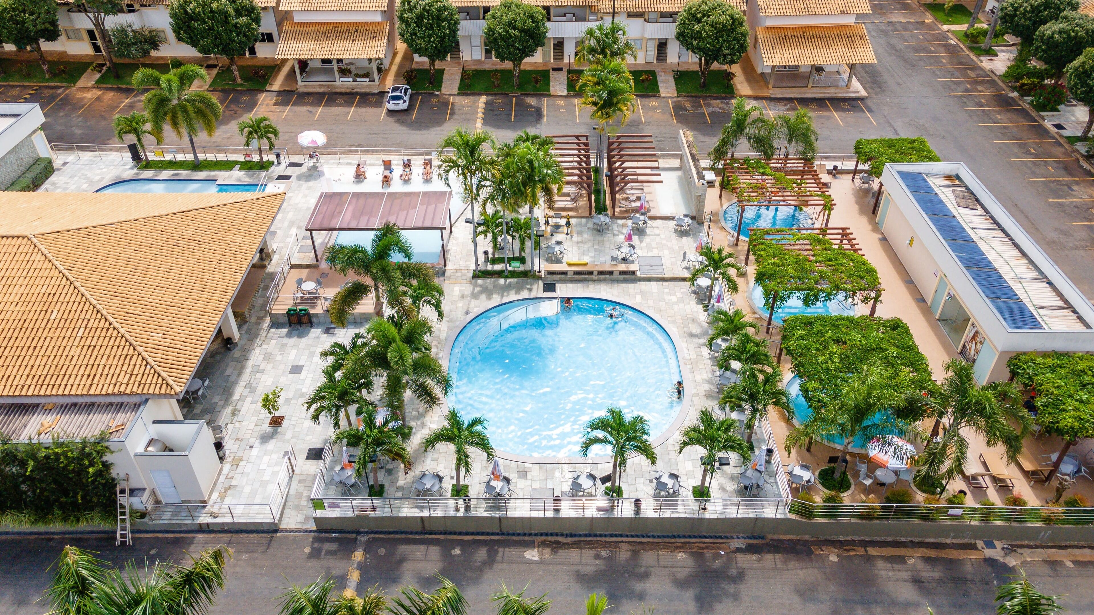An aerial view of a resort with a central swimming pool surrounded by palm trees, lounge chairs, and a covered patio area.