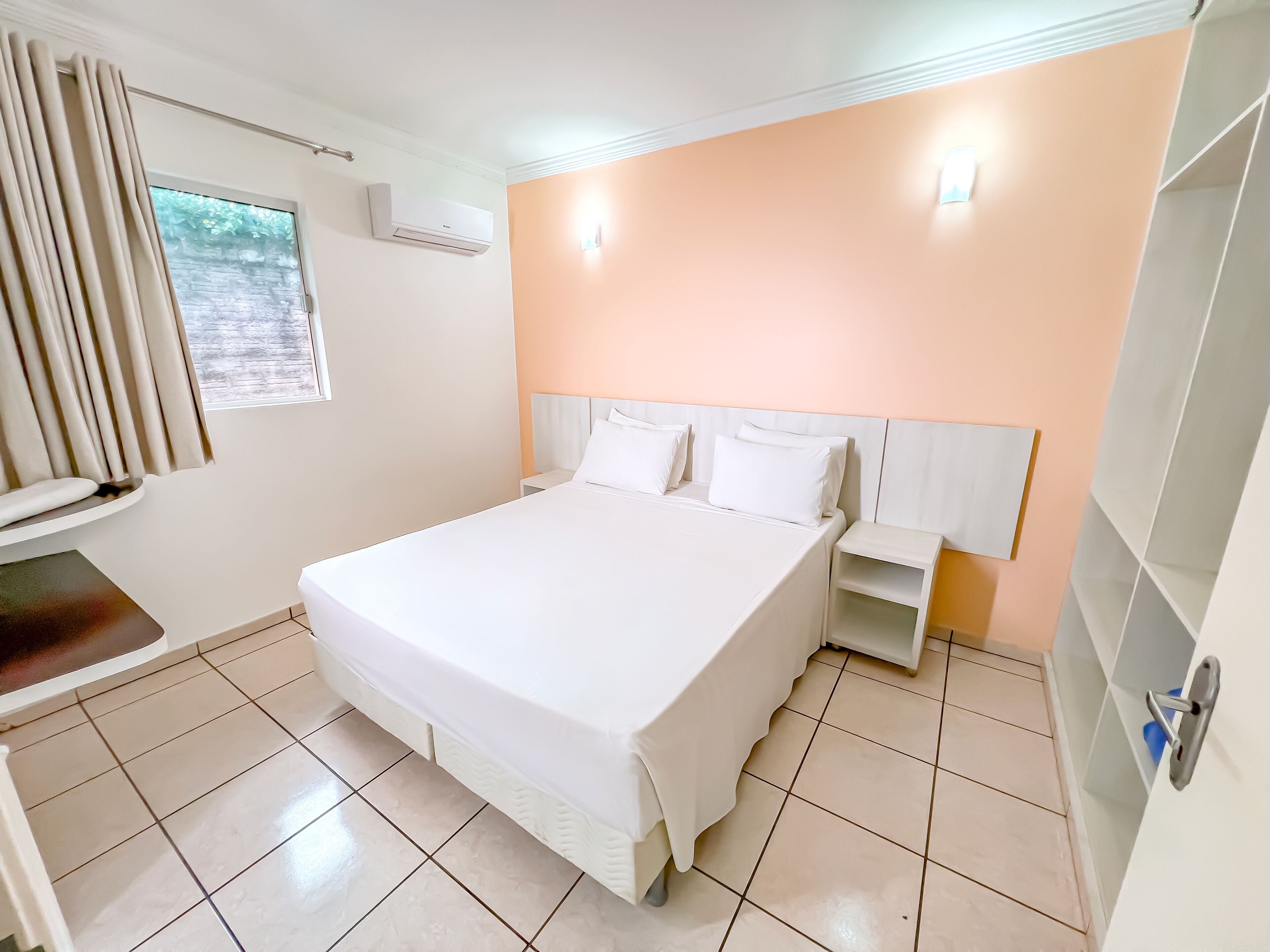 A hotel room with a large bed, a bedside table, a nightstand, a window with curtains, an air conditioning unit, and a wall-mounted light fixture.