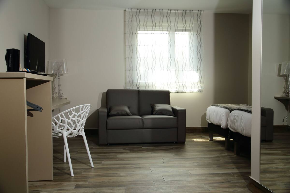 Une chambre moderne avec un canapé gris, une chaise blanche, un lit et un bureau équipé d’un ordinateur et d’une lampe.