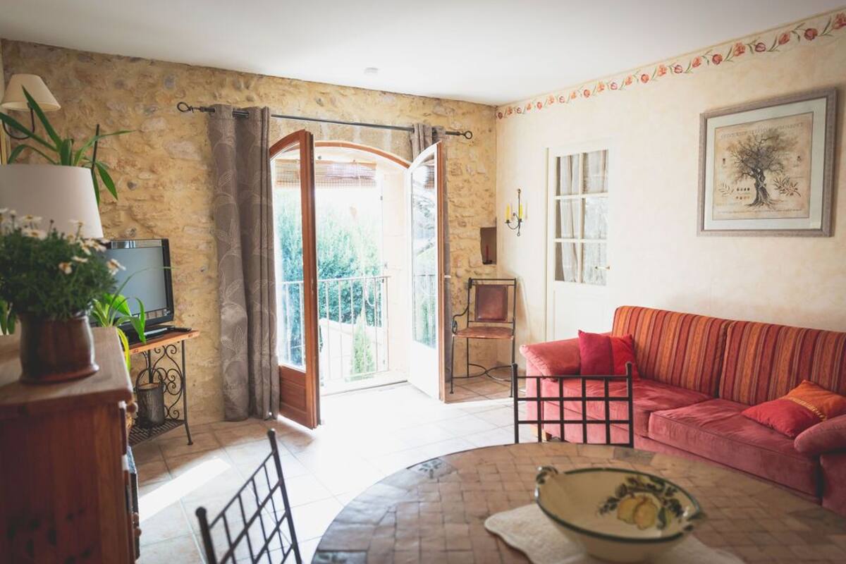 Un salon chaleureux avec un canapé rouge, une table ronde et un téléviseur posé sur un meuble en bois. Il y a un balcon avec vue sur la verdure.
