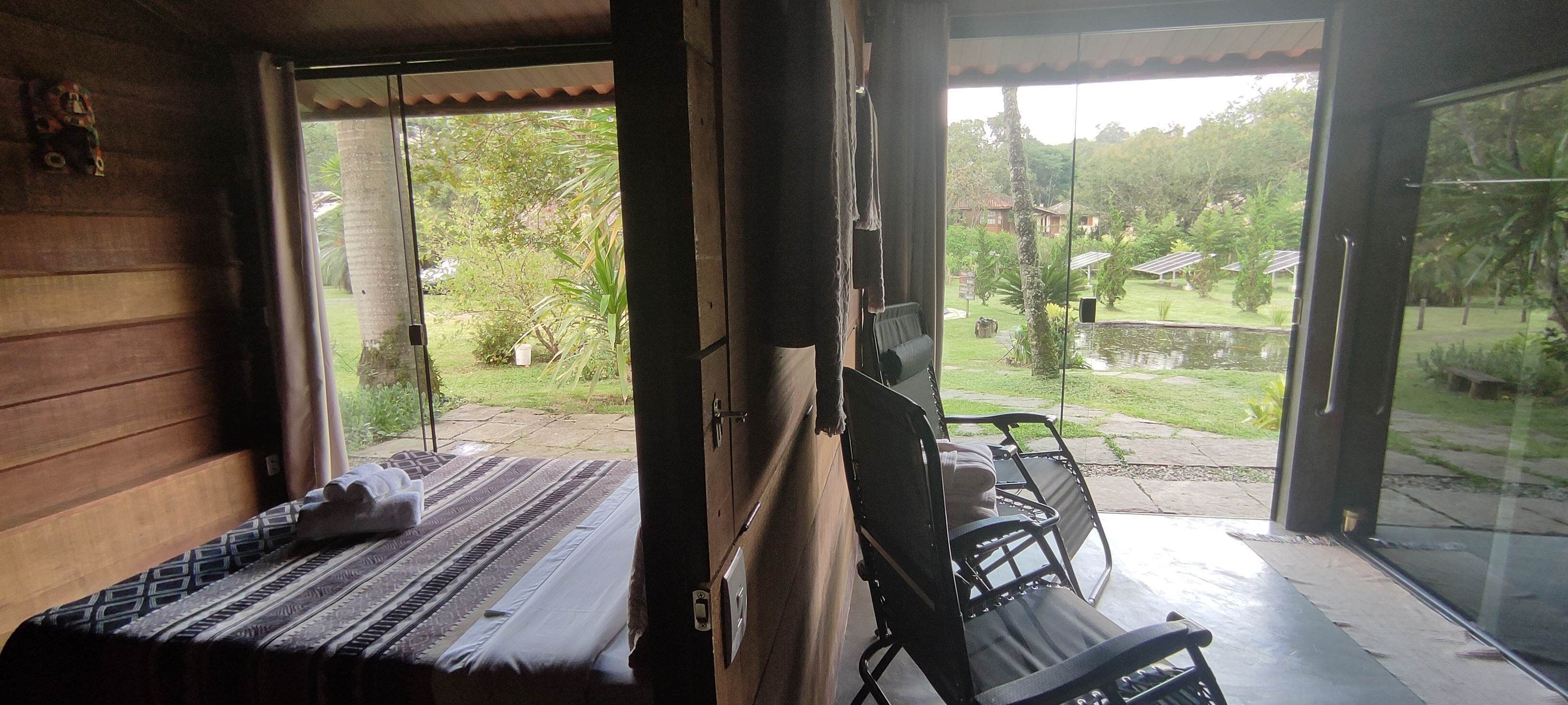 Quarto com cama, uma cadeira e vista para um jardim através de uma porta de vidro.
