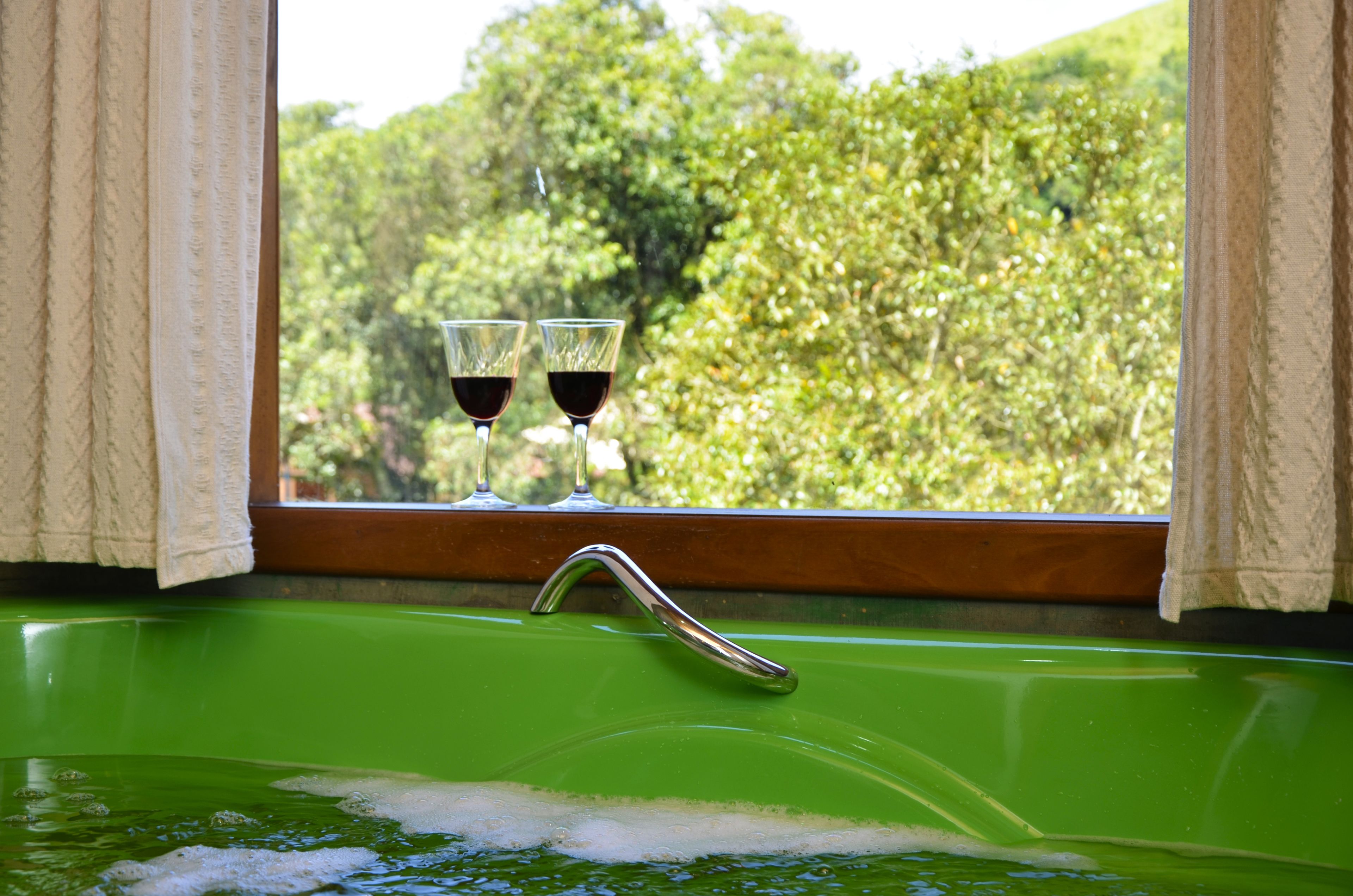 Une baignoire verte avec deux verres à vin posés sur un rebord de fenêtre, une fenêtre avec des rideaux et une vue sur des arbres.