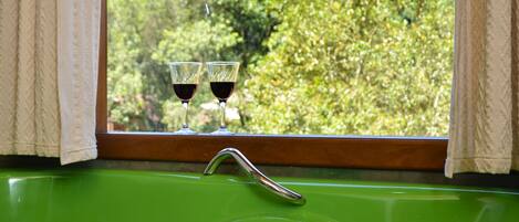 Une baignoire verte avec deux verres à vin posés sur un rebord de fenêtre, une fenêtre avec des rideaux et une vue sur des arbres.