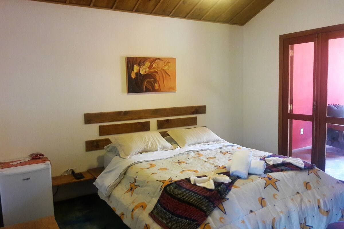 Un dormitori amb un panell de fusta al capçal del llit, un llit amb un edredó estampat, un quadre a la paret i una porta que dóna a l'exterior.