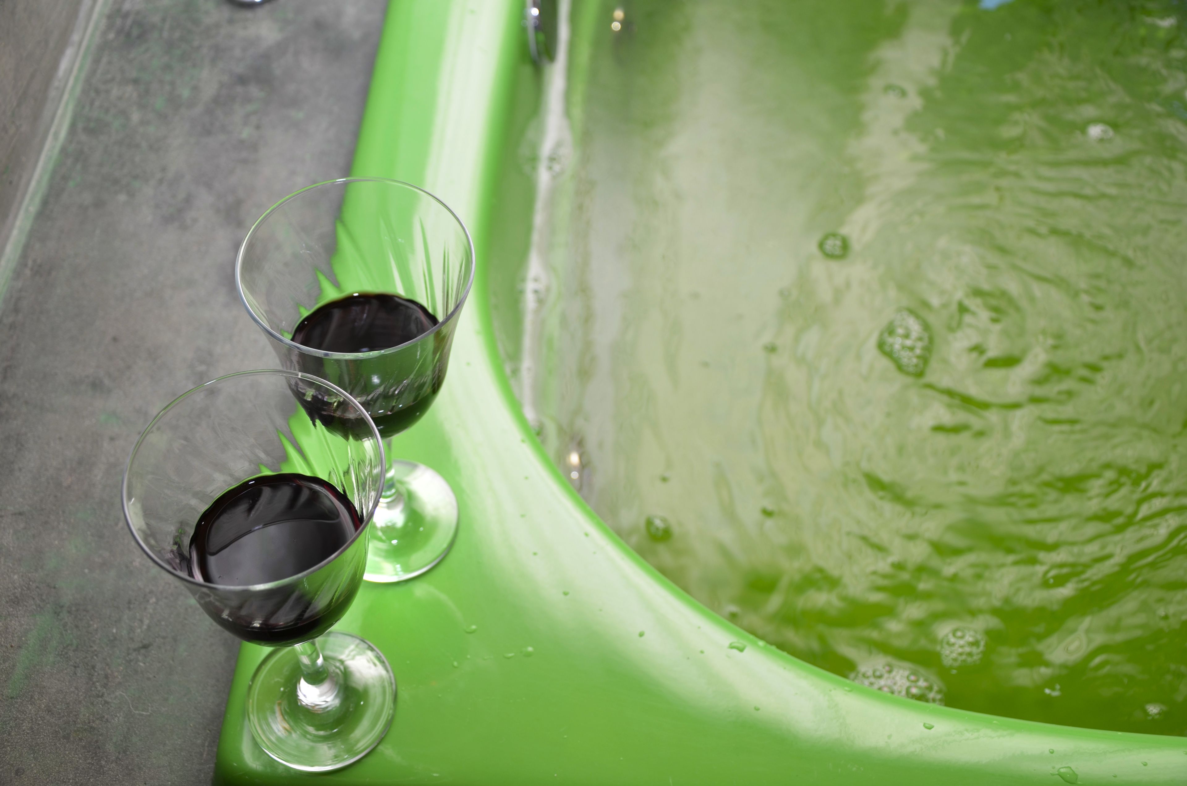 Two glasses of red wine on a green surface next to a bathtub filled with water.