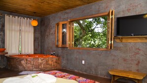Ein Zimmer mit einer Steinbadewanne, einer Holzbank, einem großen Fenster mit Blick auf Bäume und einem an der Wand befestigten Fernseher.