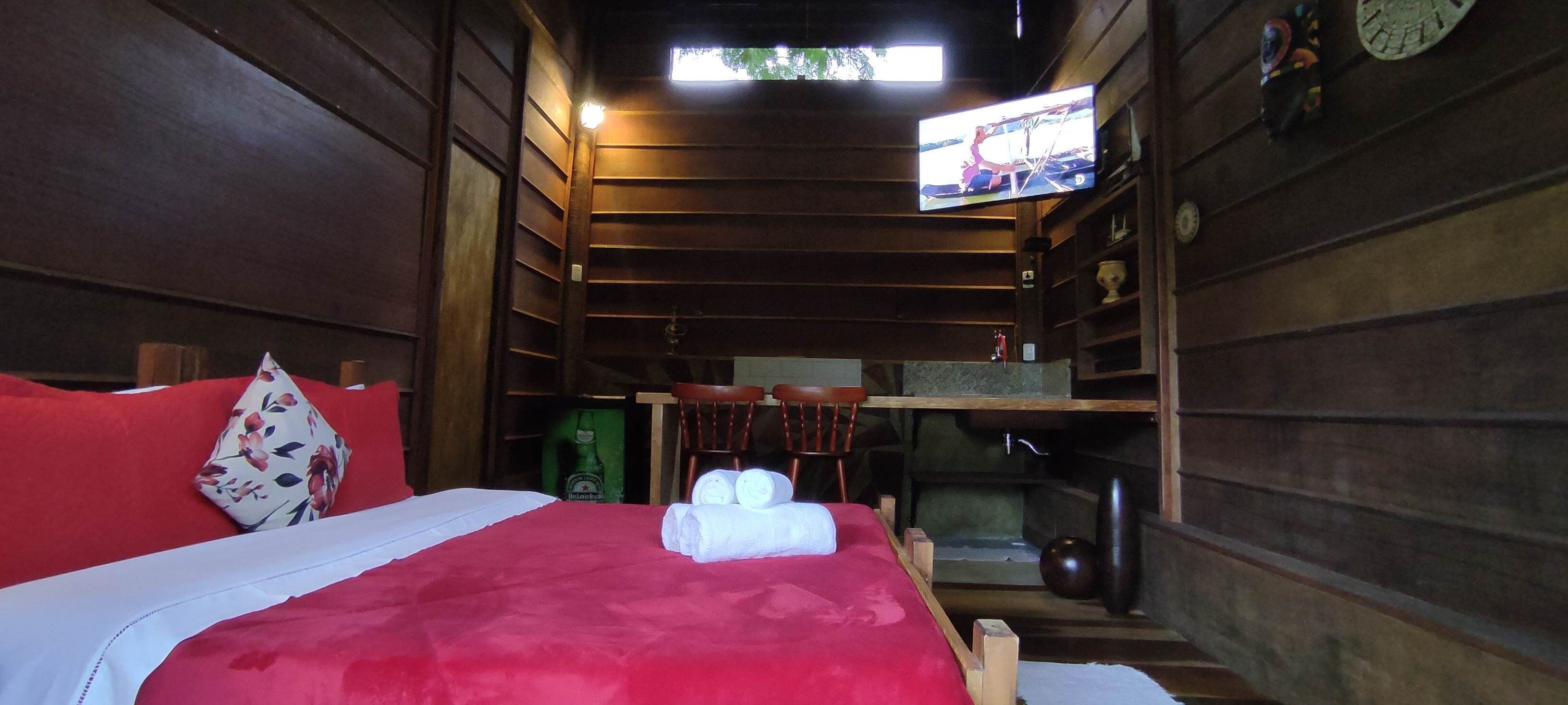 A wooden cabin room with a bed, a television mounted on the wall, and a small shelf with a clock and decorative items.