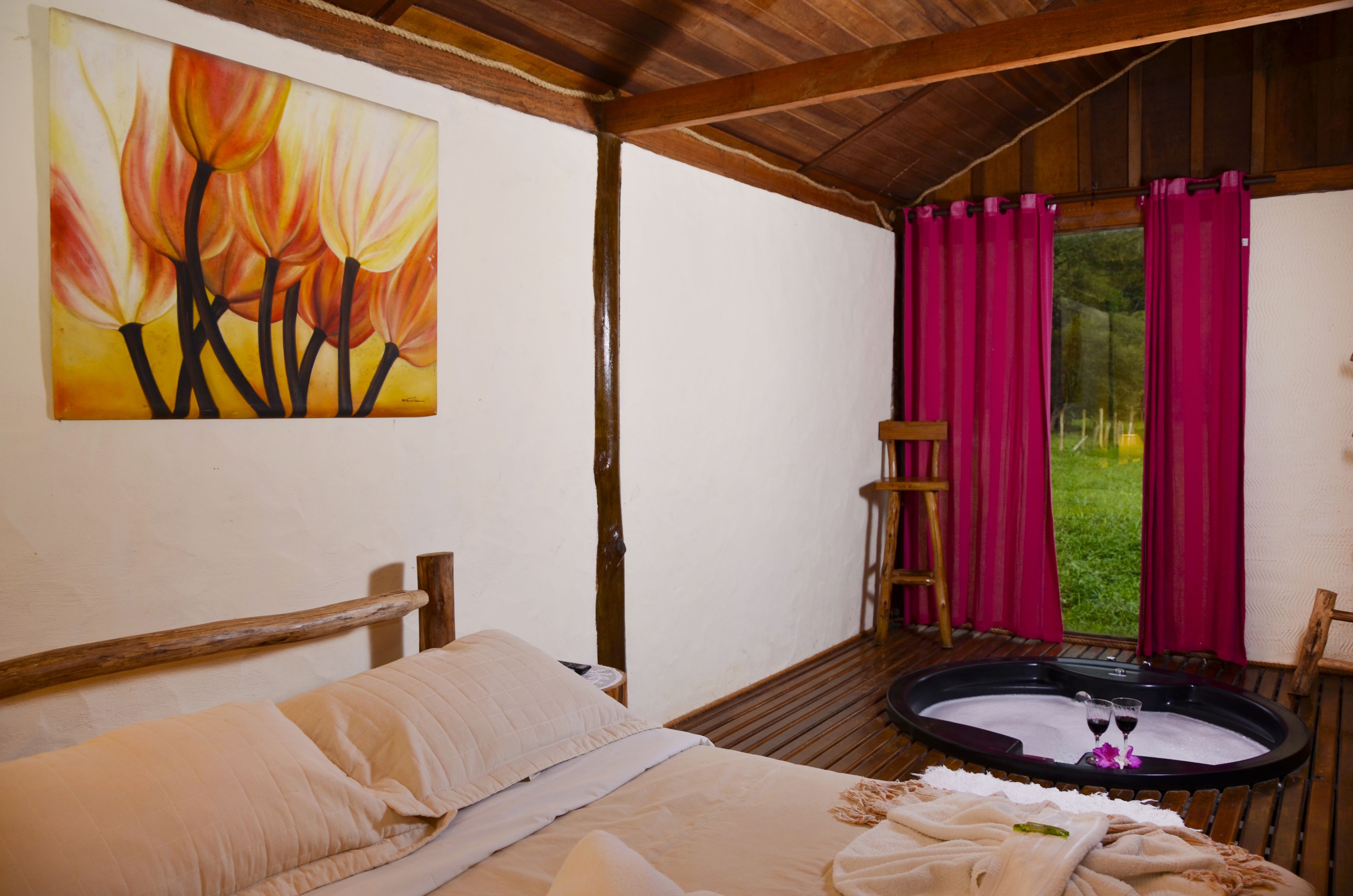 A room with a bed, a painting, a wooden chair, a small table with a glass, and a view of greenery through a window with curtains.
