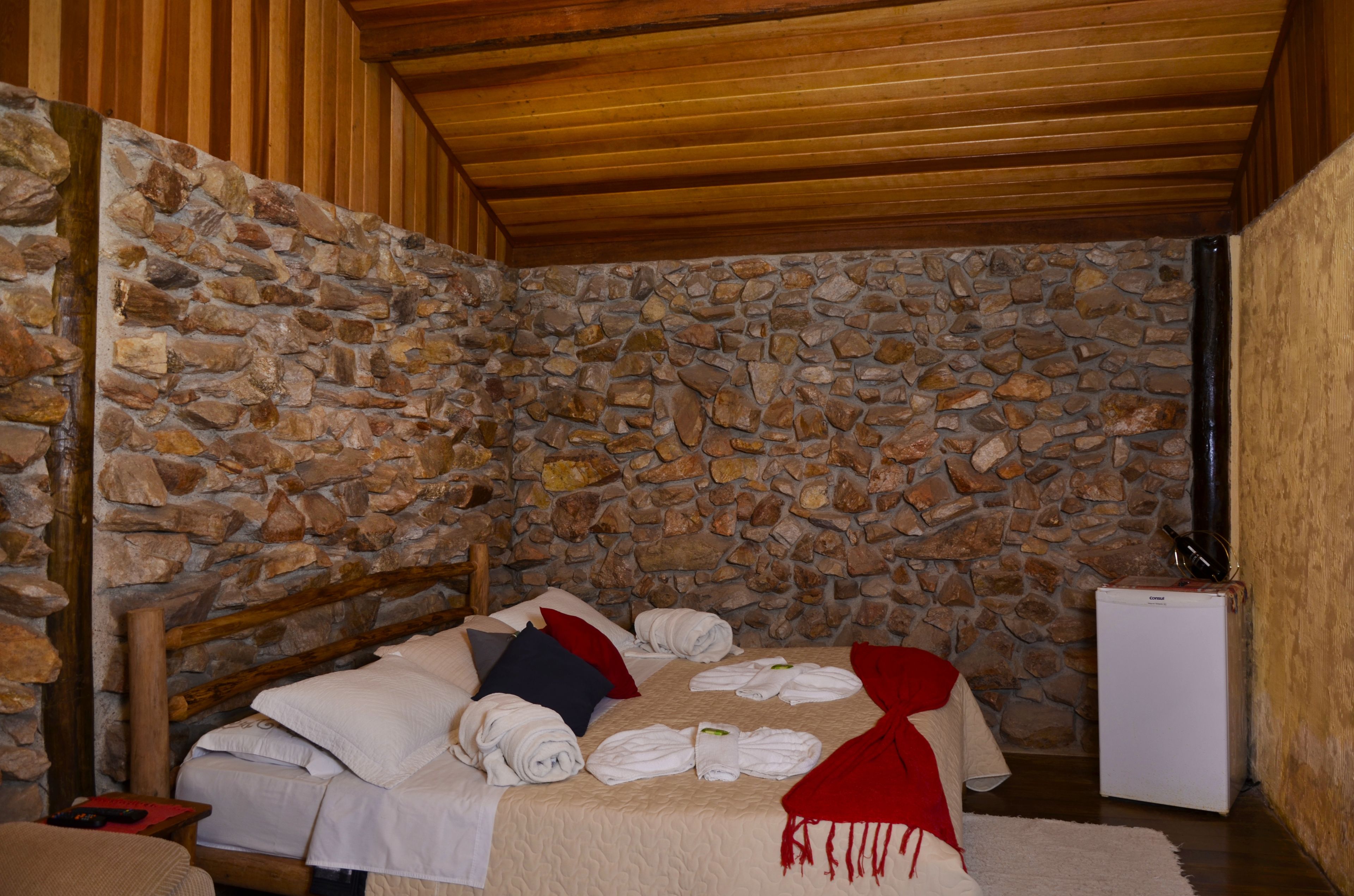 A room with a stone wall, wooden ceiling, a bed with towels, a small white refrigerator, and a wooden nightstand.