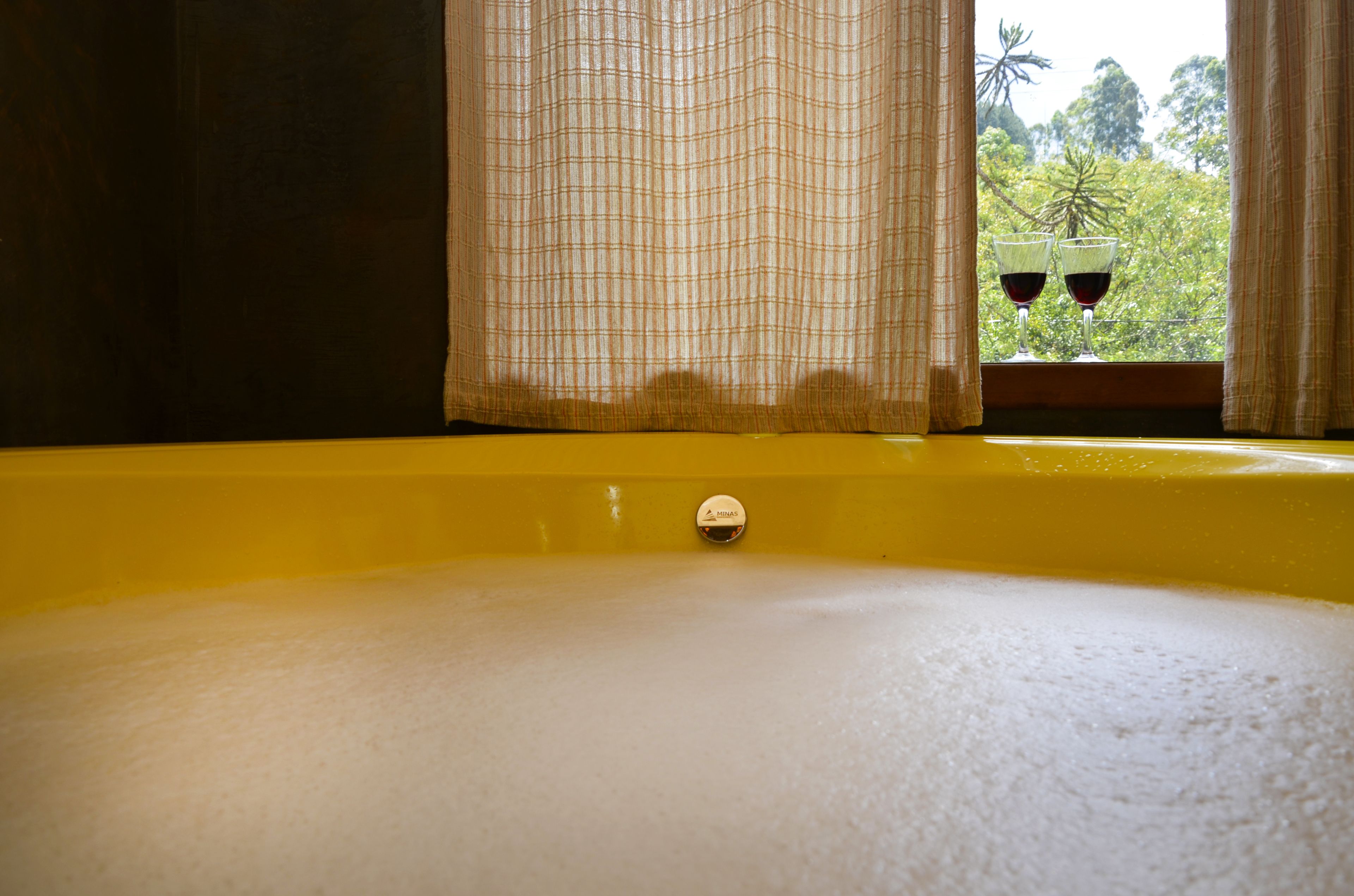 A bathtub with a yellow interior, a window with curtains, and two glasses of red wine on the windowsill.