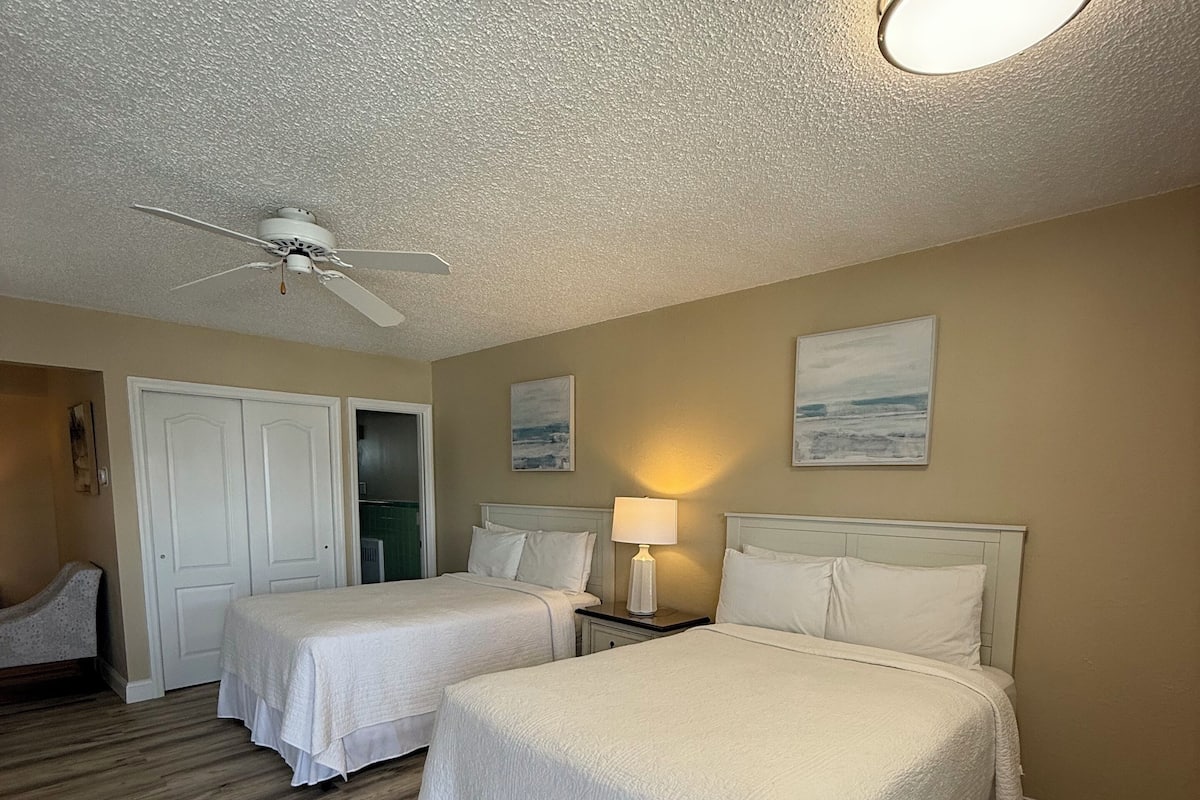 A hotel room with two beds, a ceiling fan, and a painting on the wall.