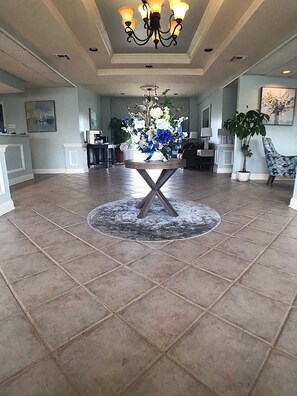 A spacious lobby with a central round table, a chandelier, and potted plants. - Point Pleasant Manor (Point Pleasant Beach)