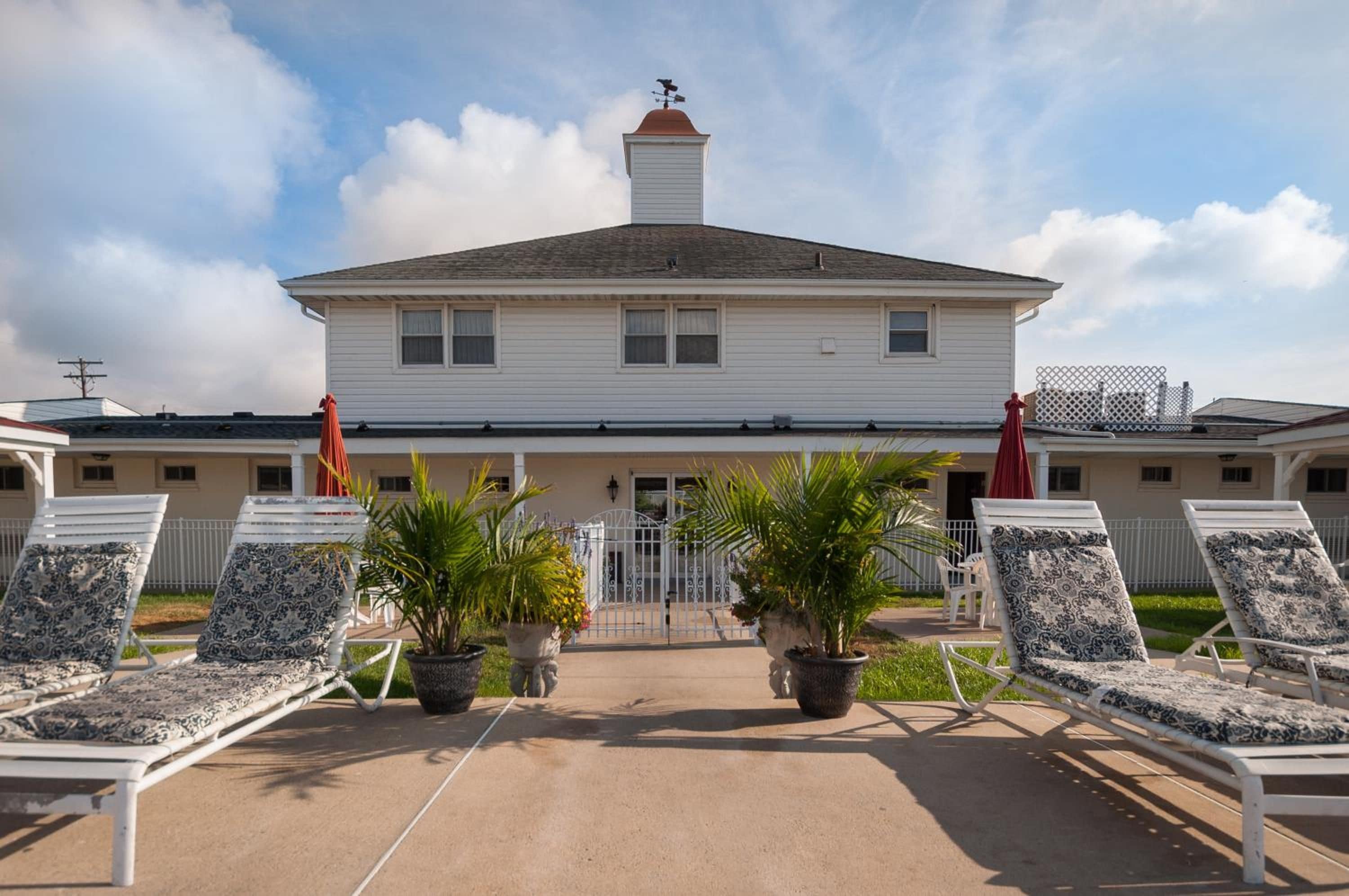 A two-story building with a central entrance, a covered patio area with lounge chairs and potted plants, and a clear sky.