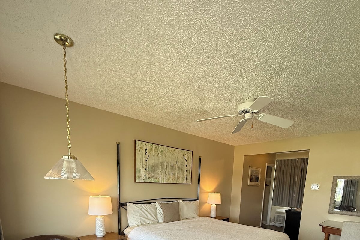 A hotel room with a bed, two bedside lamps, a ceiling fan, a wooden floor, and a round table with a chair.