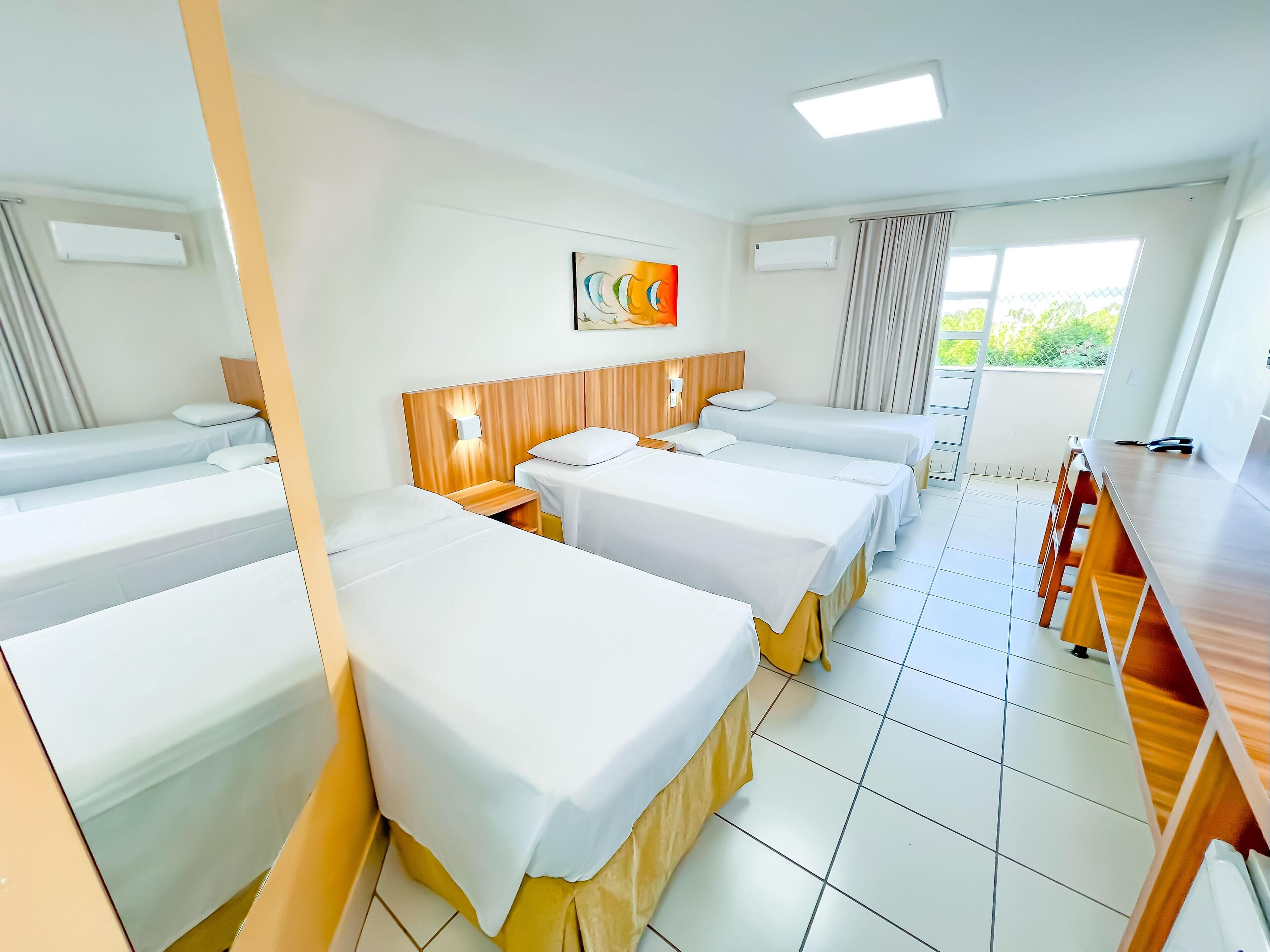Une chambre d’hôtel avec quatre lits simples, une tête de lit en bois, une petite table et une fenêtre avec des rideaux.