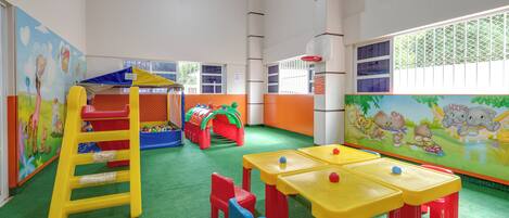 A brightly colored indoor play area with a climbing structure, tables, chairs, and a ball pit.