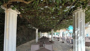 An outdoor cafe with wicker tables and chairs, surrounded by white columns and covered by a leafy canopy. - DiRoma Exclusive - Direto com o Grupo DiRoma - acesso ao Acqua Park com café da manhã (Caldas Novas)