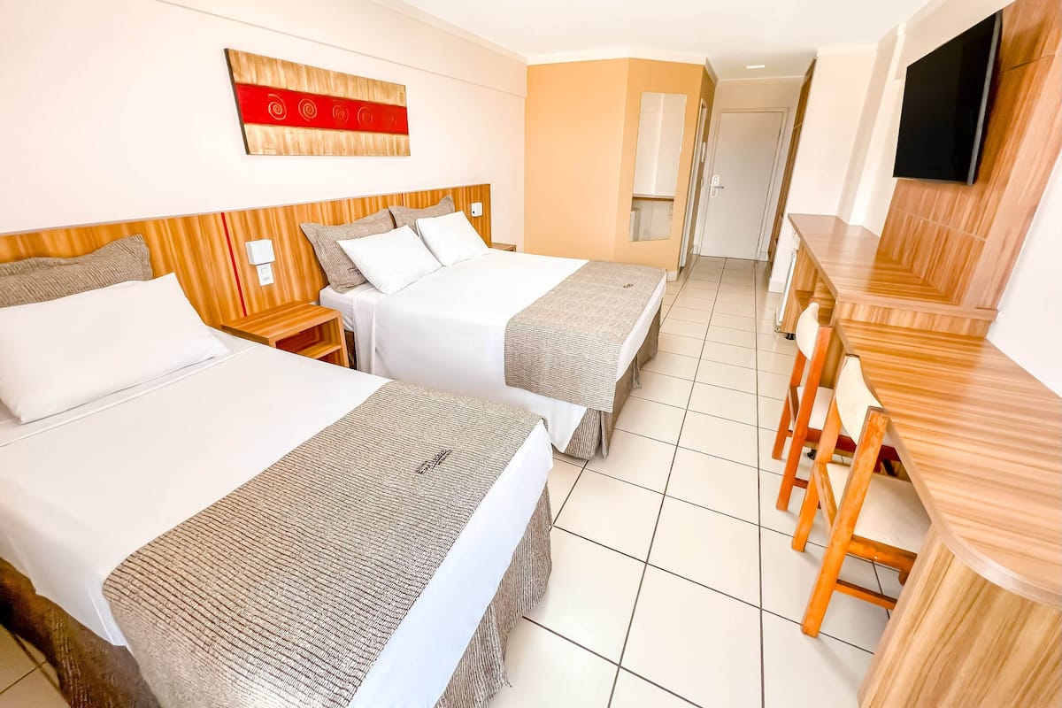 A hotel room with two beds, a wooden headboard, a flat-screen TV, a wooden dining table, and a small TV in the corner.