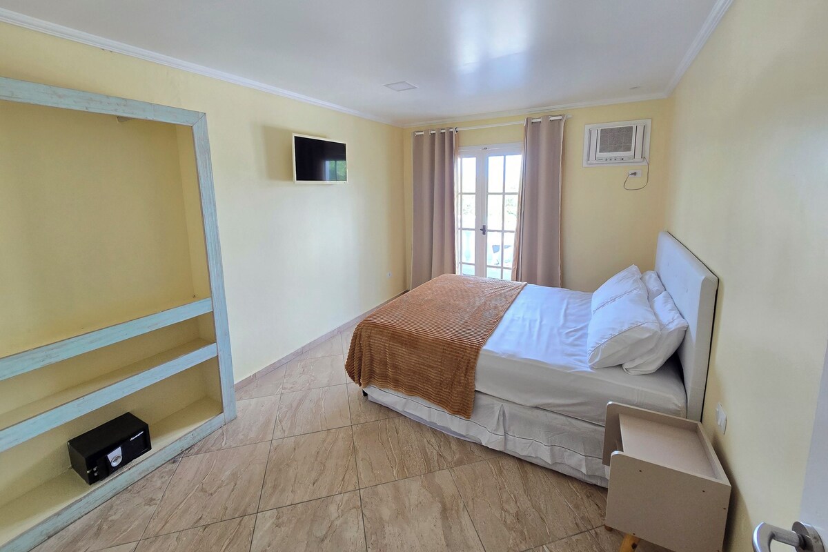 A bedroom with a bed, a TV mounted on the wall, a window with curtains, and a small shelf with a box.