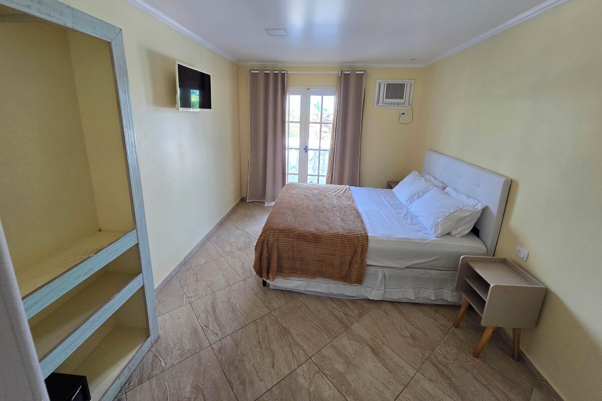 A bedroom with a bed, a small bedside table, a window with curtains, and a wall-mounted air conditioning unit.