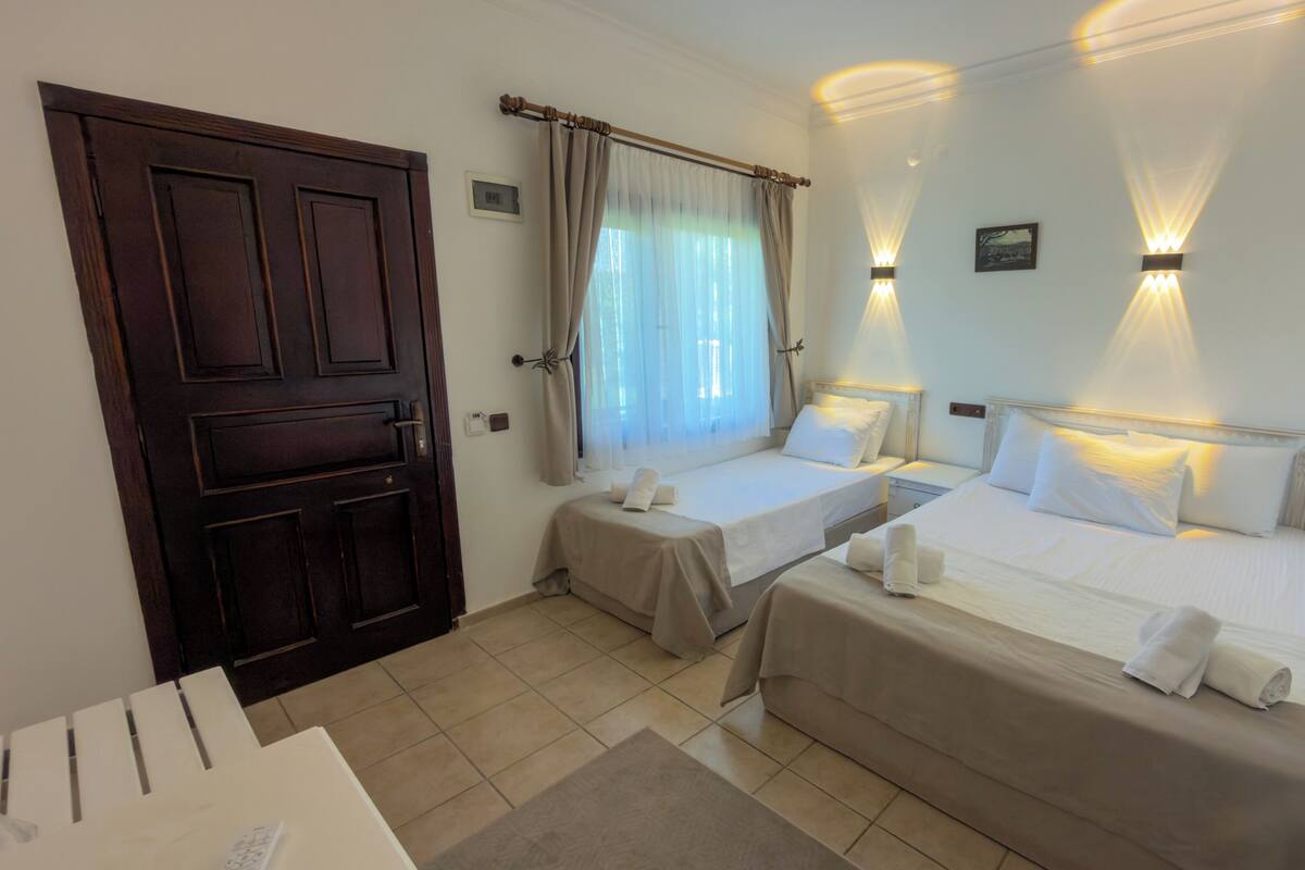 Quarto de hotel com duas camas de solteiro, porta de madeira, janela com cortinas e luminárias fixadas na parede.