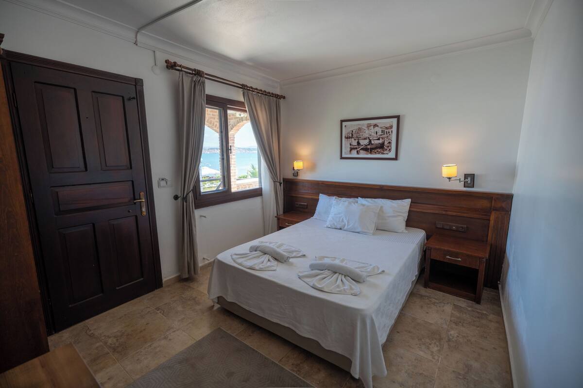 Quarto de hotel com uma cama, uma mesa de cabeceira de madeira, uma luminária fixada na parede e uma porta com cortinas.