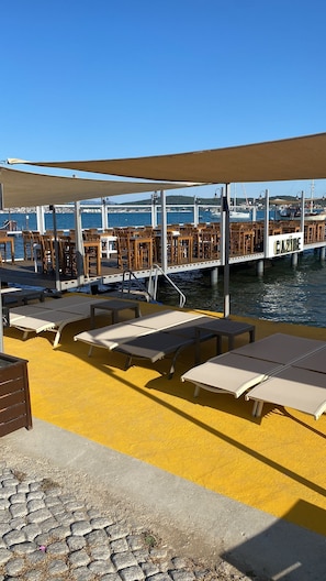 Un restaurante frente al agua con terraza, que incluye sillones de sol y mesas, además de una zona cubierta.