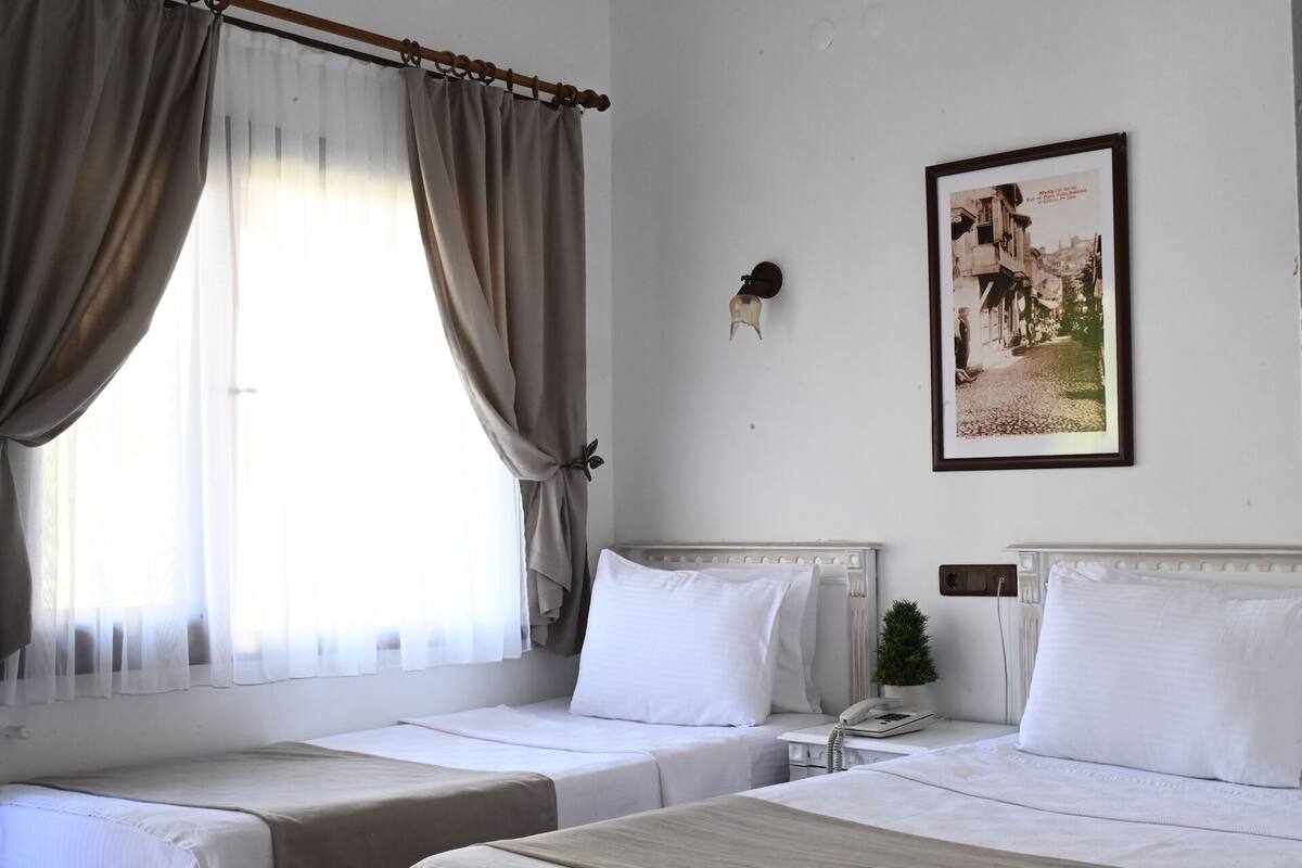 Quarto de hotel com duas camas, janela com cortinas, telefone fixado na parede e um quadro emoldurado.