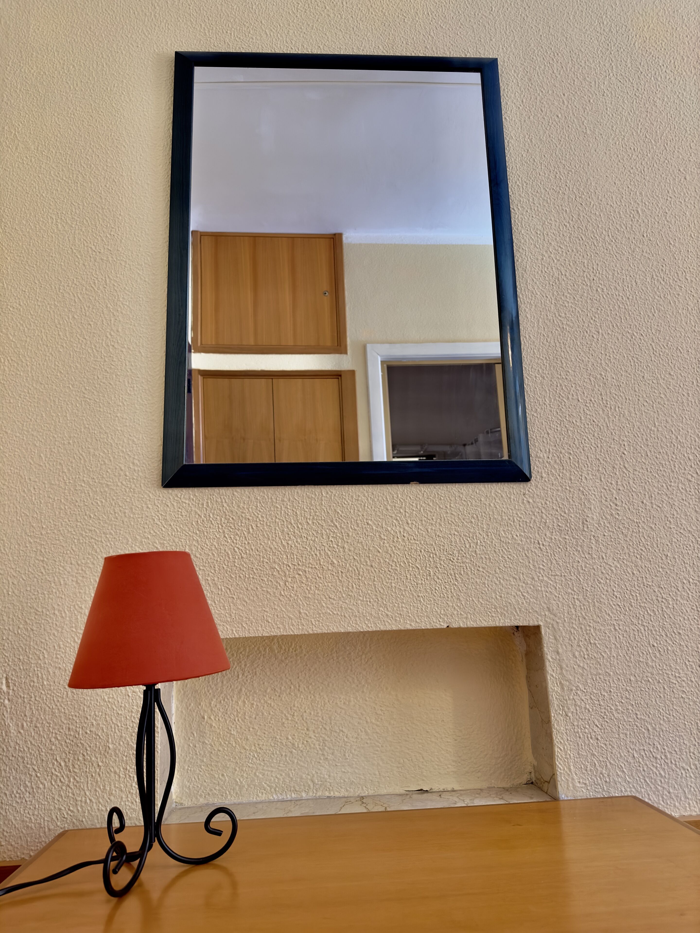 A mirror with a black frame on a textured wall, a red lampshade, and a wooden table below.
