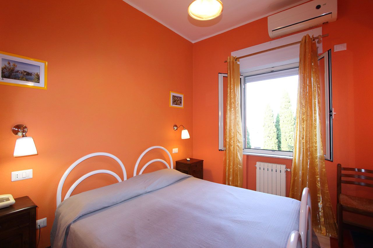 Ein Schlafzimmer mit oranger Wand, einem Bett mit weißem Kopfteil, einem Nachttisch, einer Lampe, einem Sessel und einem Fenster mit Vorhängen.