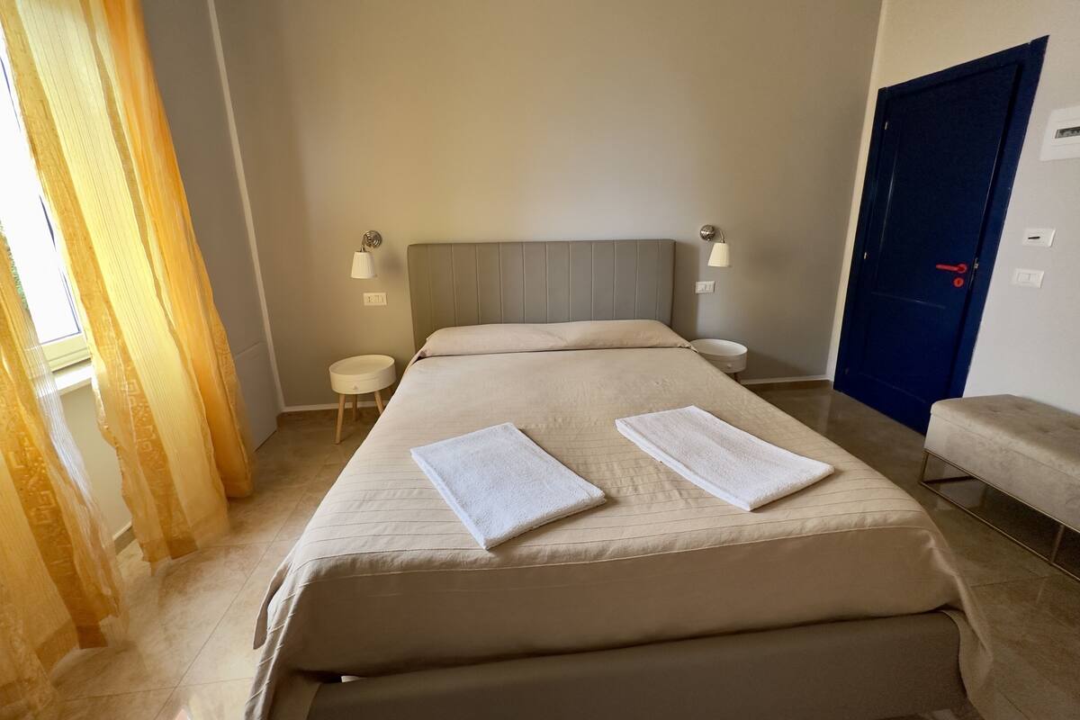 A neatly made bed with two towels, a bedside table, a lamp, and a blue door.