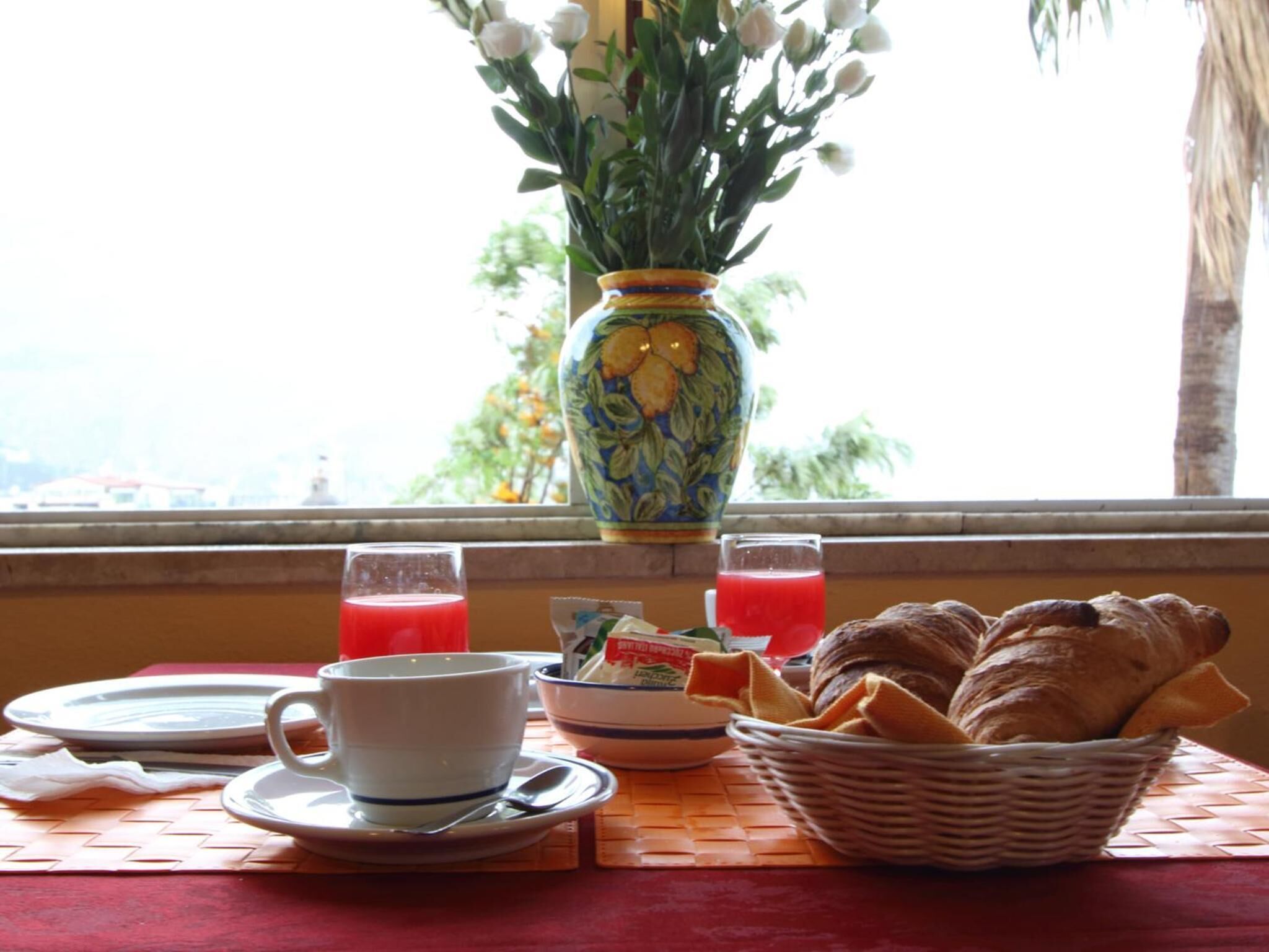 A breakfast setup with a vase of flowers, two glasses of red juice, a bowl of cereal, a cup, a plate, and a basket of croissants.