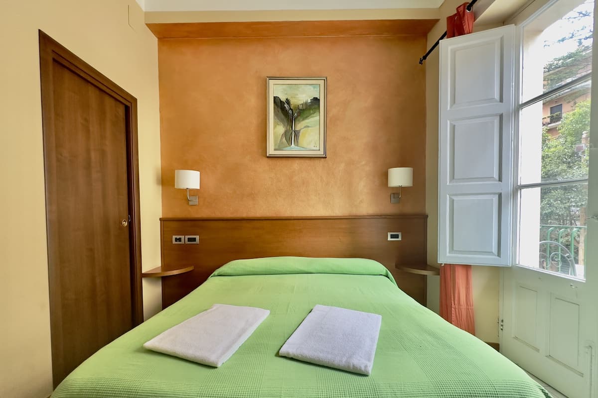 A hotel room with a green bedspread, two white towels, a painting on the wall, and a window with a view of buildings.