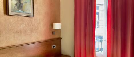 A hotel room with a green bed, red curtains, a painting on the wall, and a view of the outside.