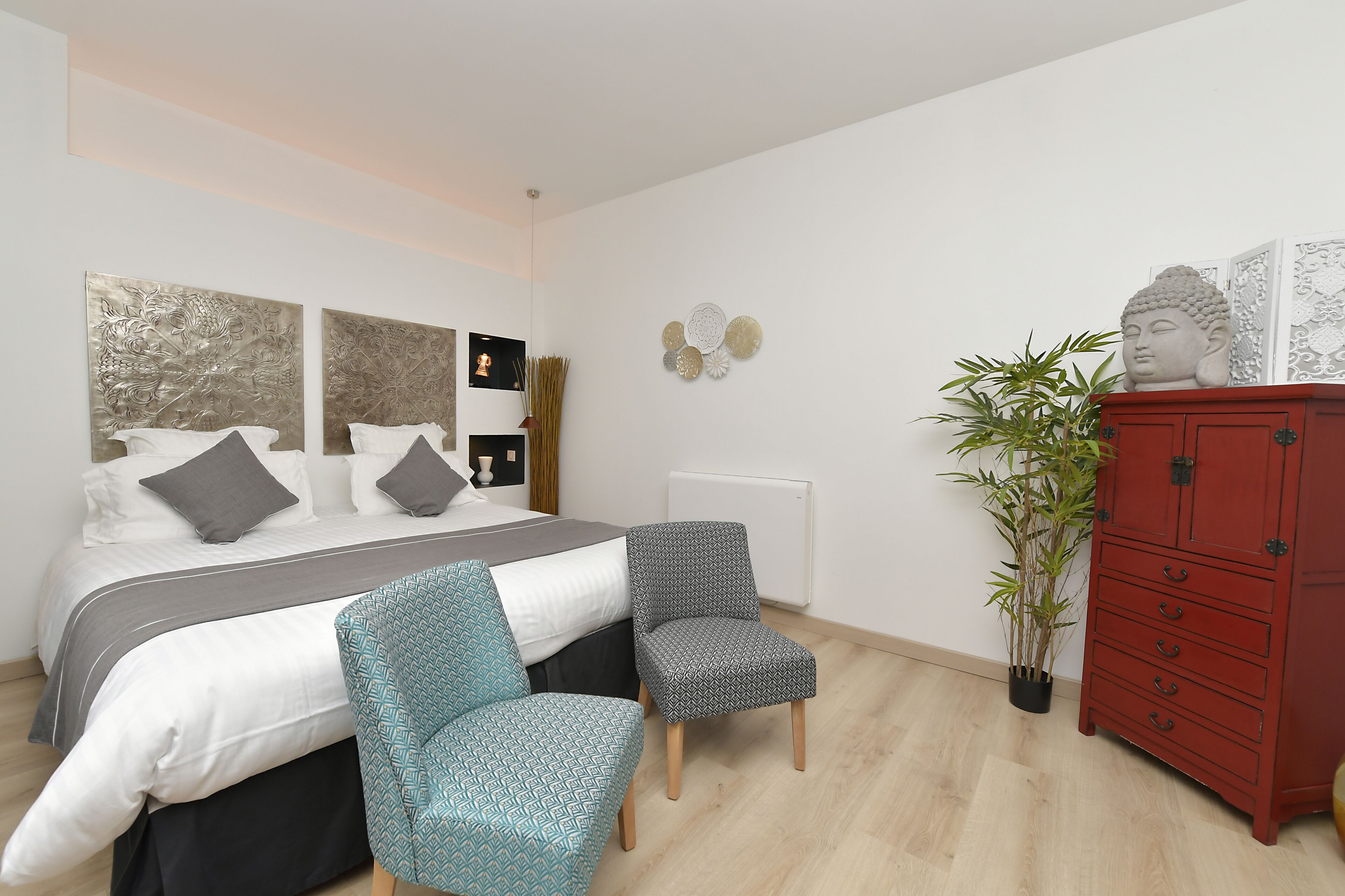 A bedroom with a bed, two chairs, a red cabinet, and a potted plant.