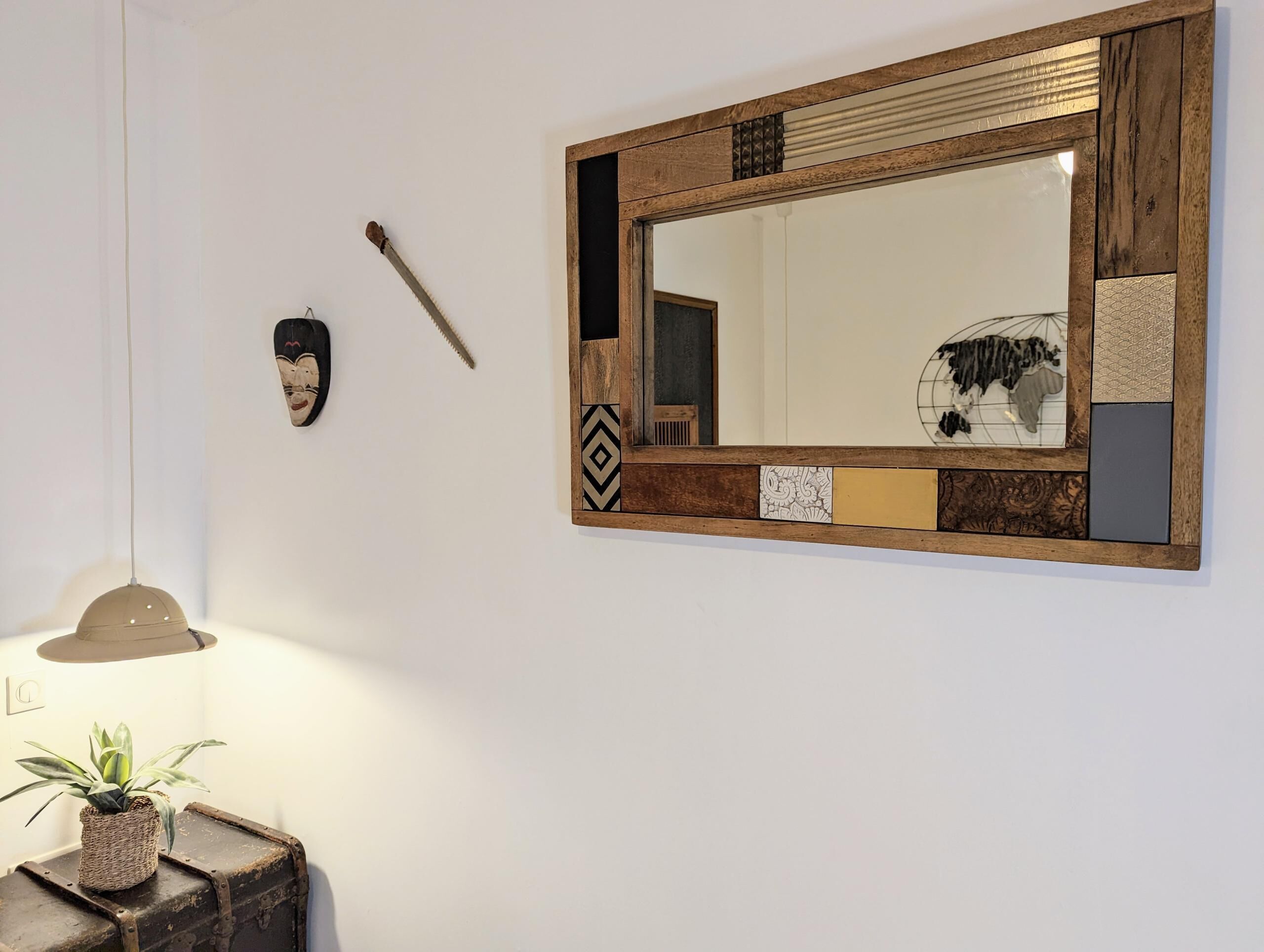 Un espejo con marco de madera y azulejos decorativos, una pequeña planta en maceta sobre una maleta antigua, y un aplique de pared.