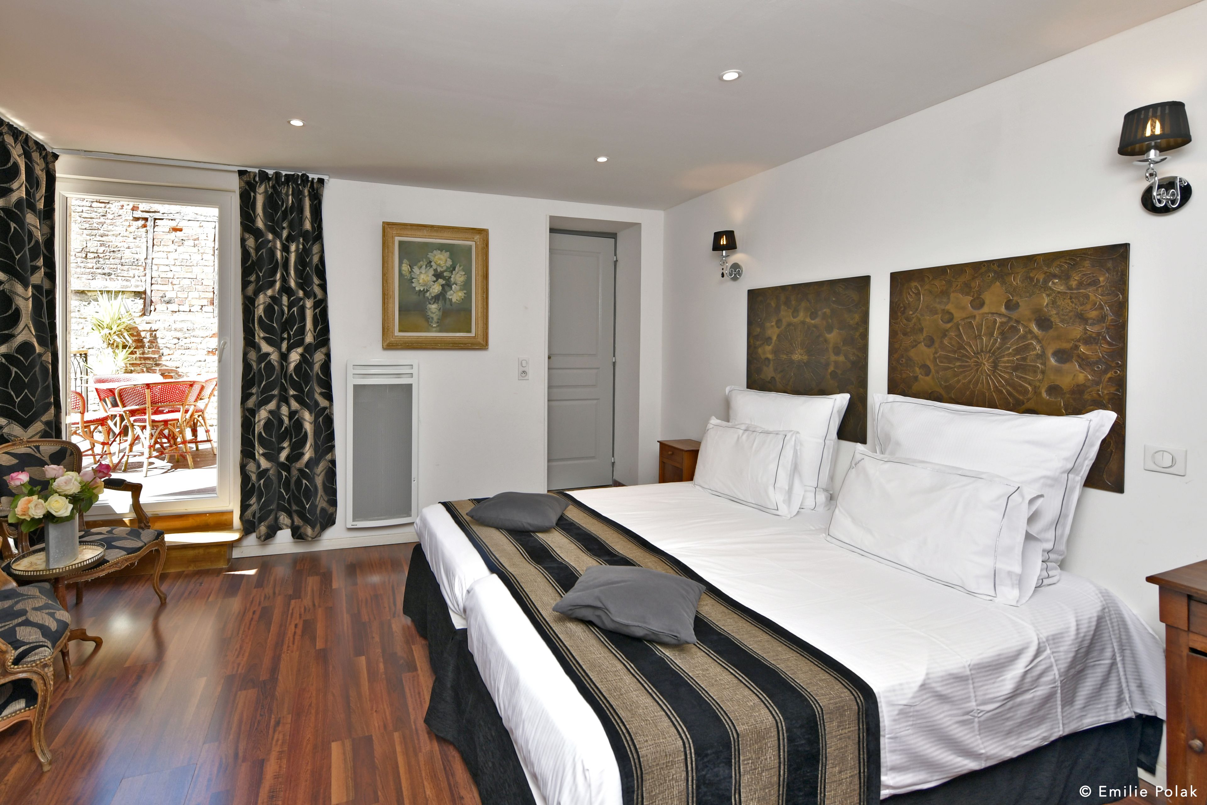 A bedroom with a large bed, a small table with chairs, a painting on the wall, and a door leading to another room.