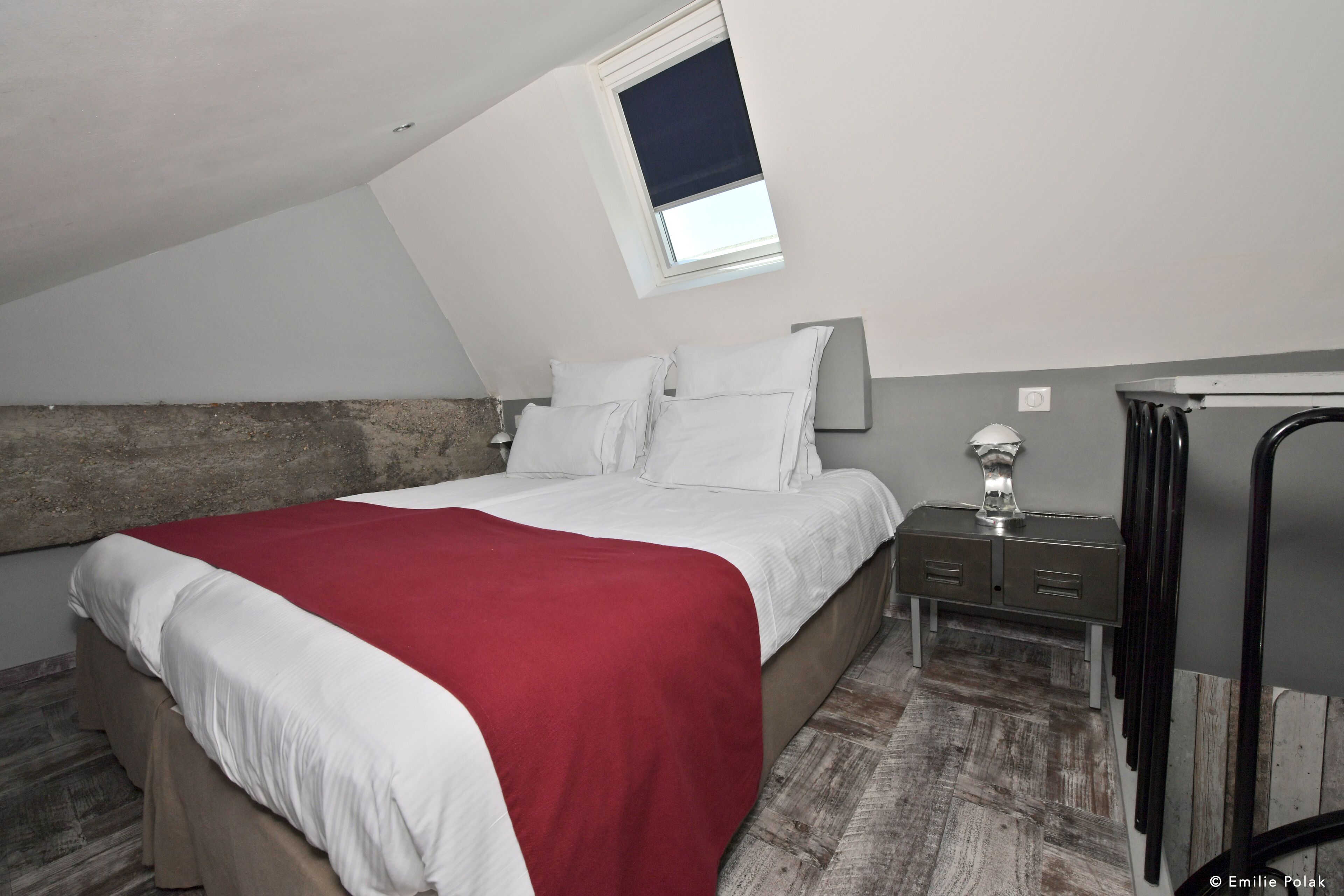 A bedroom with a sloped ceiling, a bed with white and red bedding, a nightstand with a lamp, and a small table with a vase.