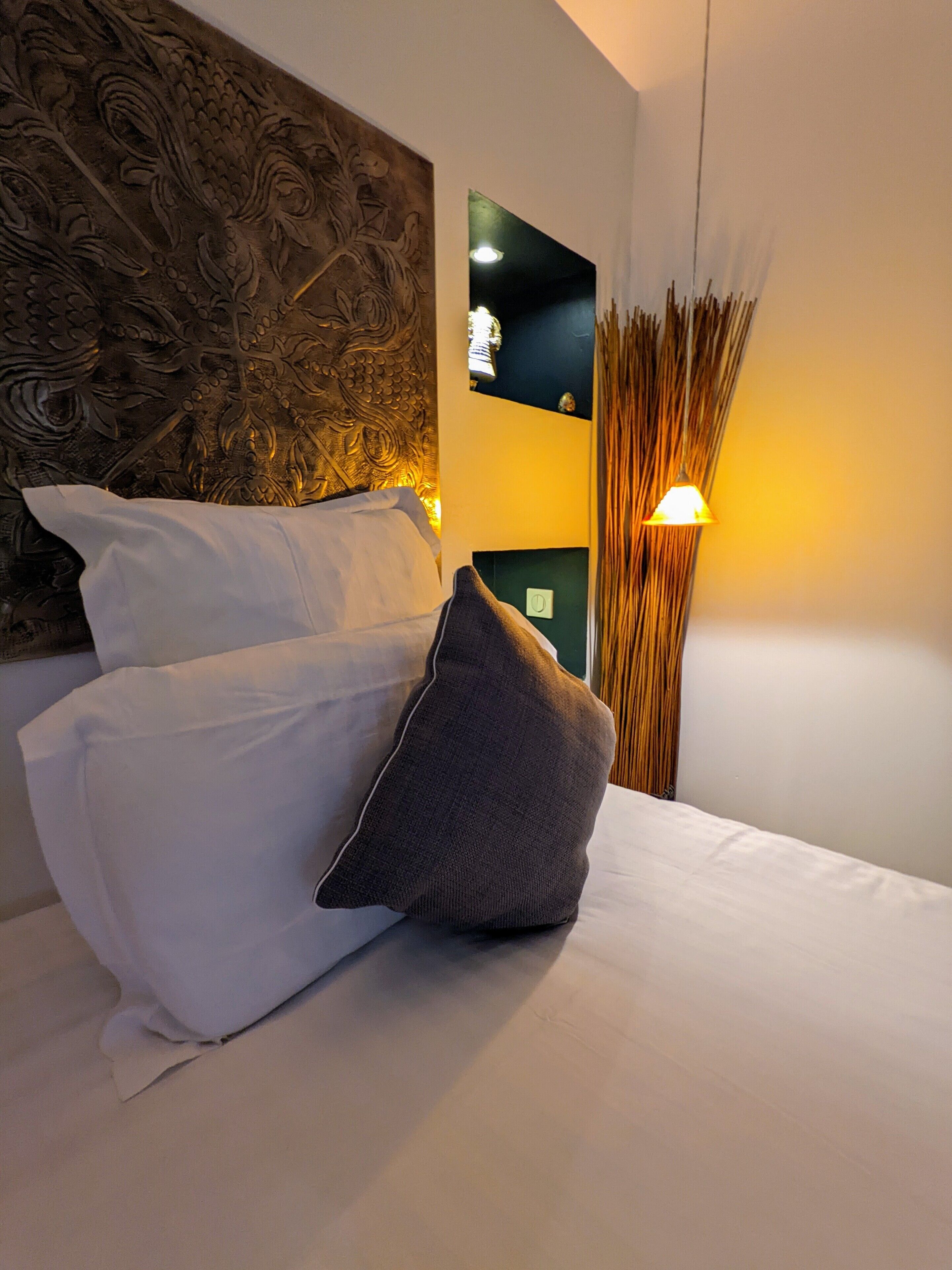 A neatly made bed with white and gray pillows, a decorative headboard, a wall-mounted light, and a tall, slender vase with dried reeds.