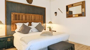 Une chambre avec une tête de lit en bois, un lit avec une literie blanche, un banc, une valise, un miroir fixé au mur et une petite plante.