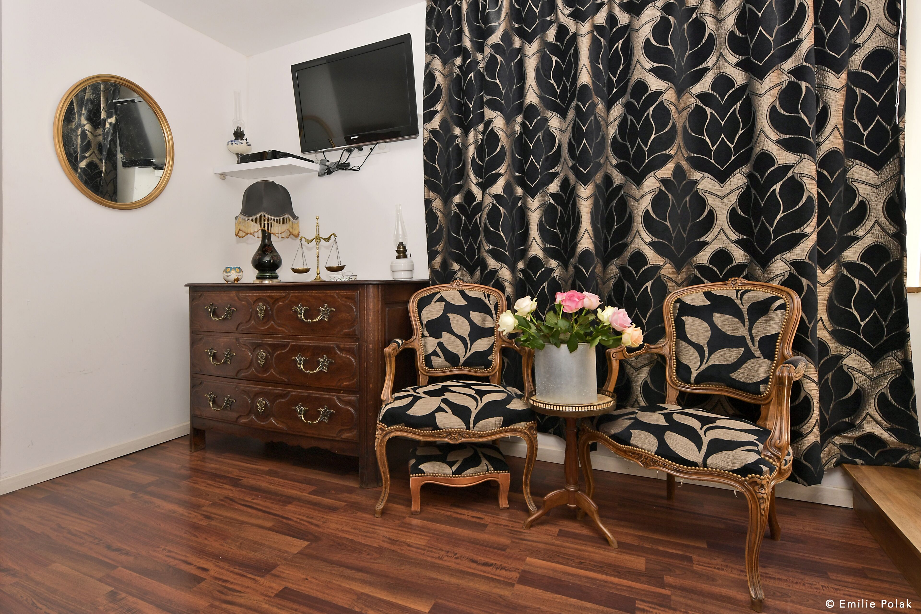 Een kamer met een houten vloer, een aan de muur bevestigde televisie, een dressoir met lades, twee gestoffeerde fauteuils met een bladmotief, een vaas met bloemen en een gordijn met patroon.