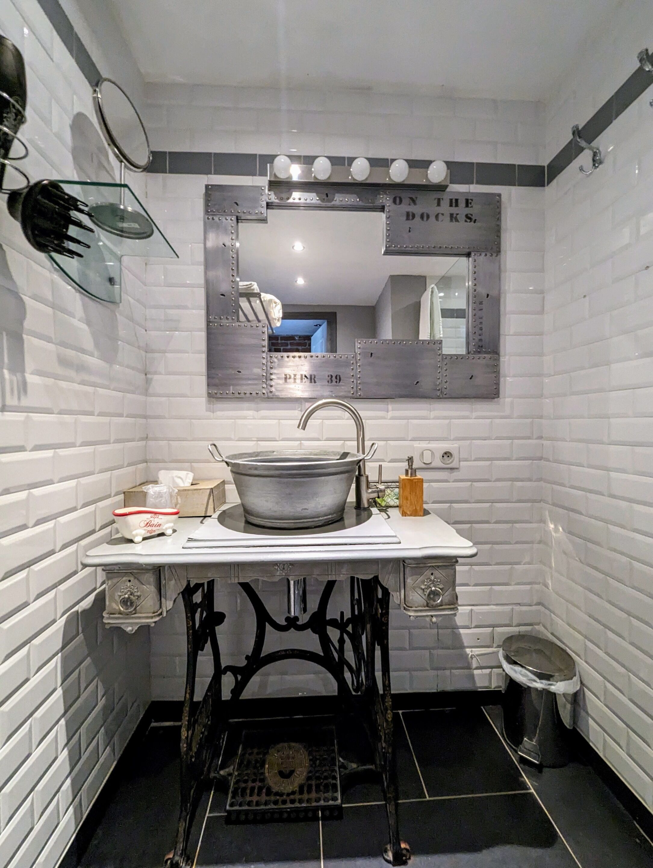 Un baño con pared de azulejos blancos, un espejo grande, un lavabo y un pequeño cubo de basura.