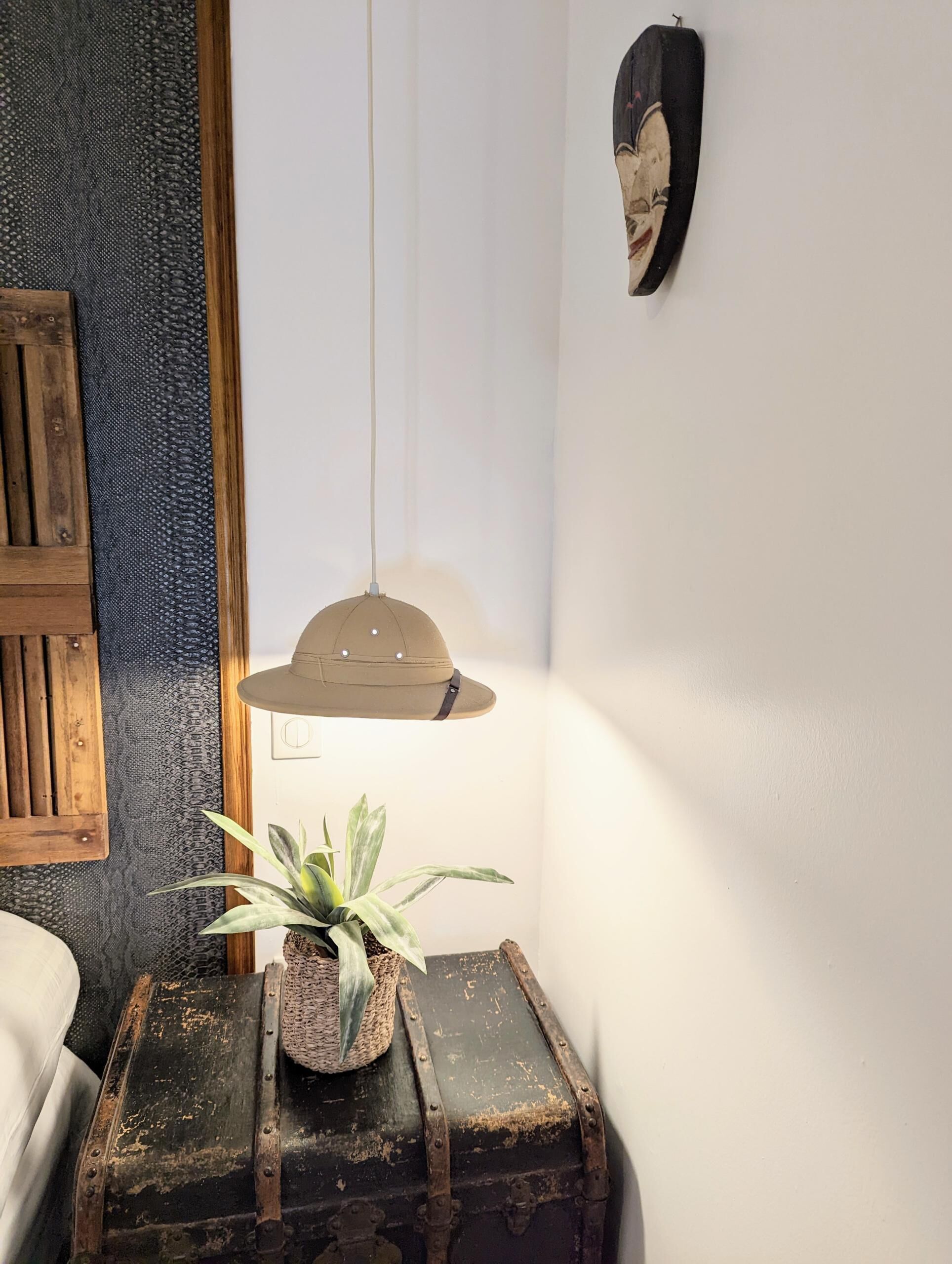 A room with a rustic wooden headboard, a hanging pendant lamp, a small table with a plant, and a vintage suitcase.