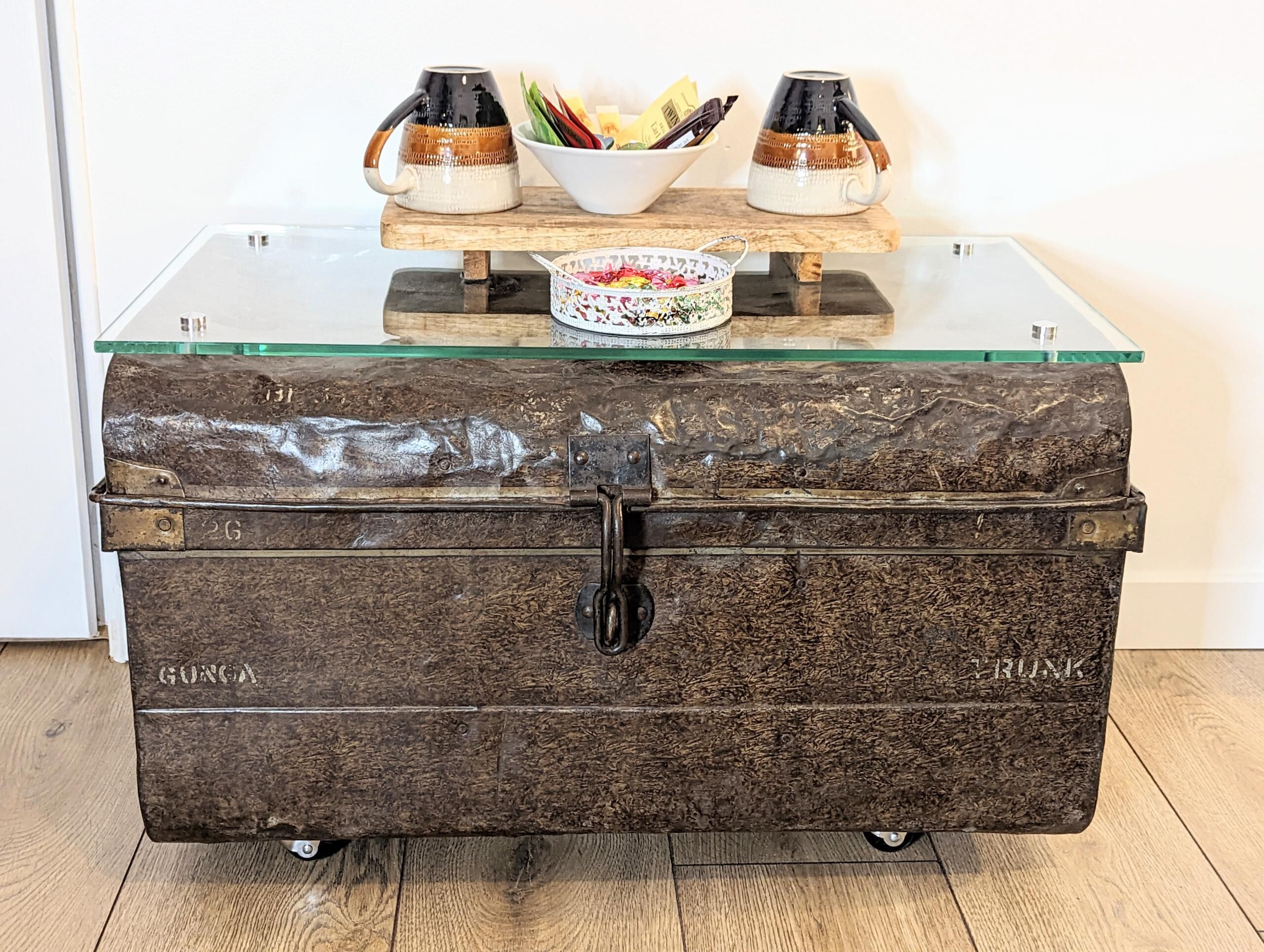 Une vieille malle en bois, dotée de poignées et de roulettes en métal, portant les inscriptions « GONGA » et « TRUNK », est posée sur une table dont le plateau est en verre, avec un bol et deux tasses.