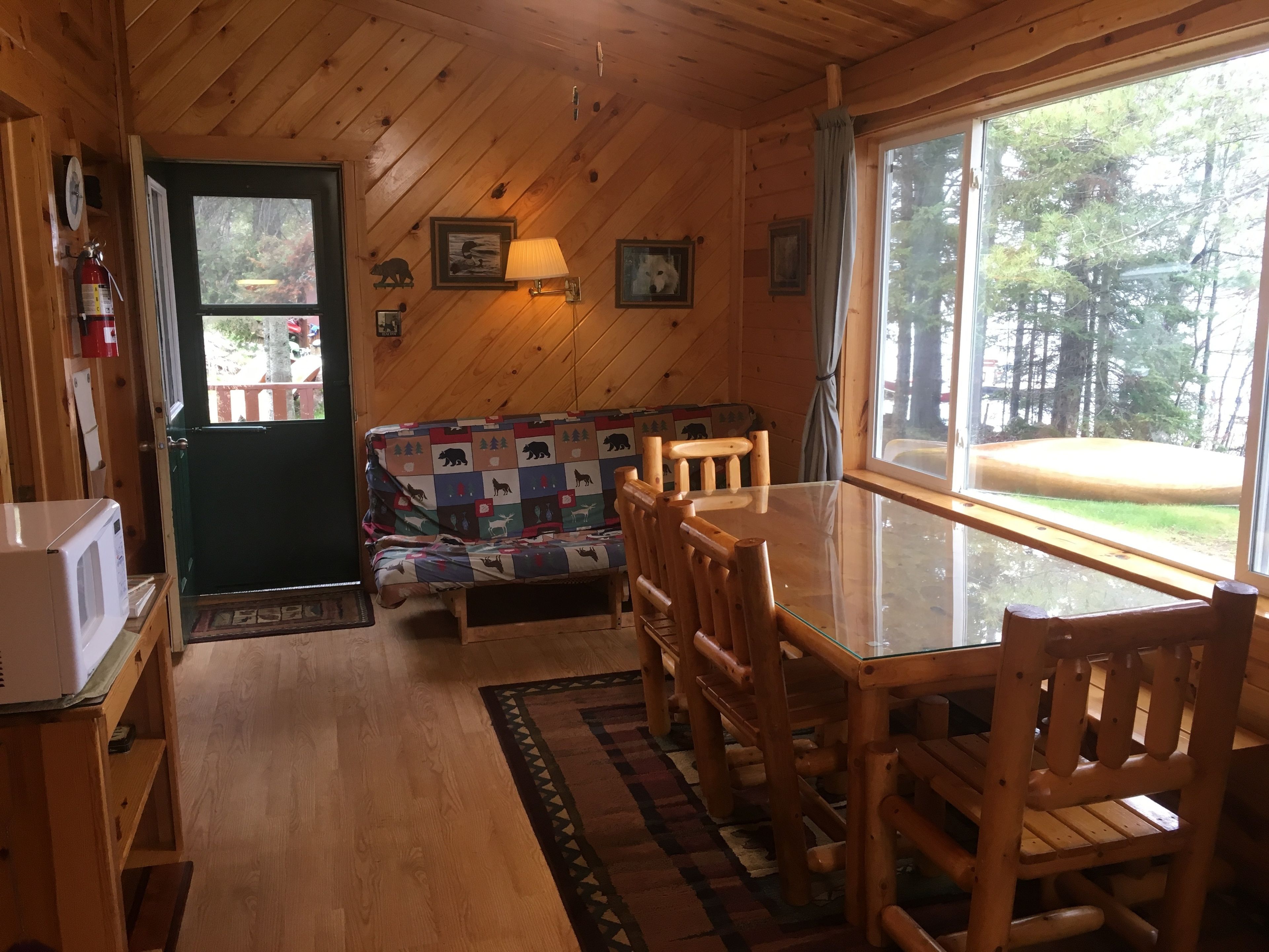 A cozy cabin interior with a wooden interior, a sofa with a bear-themed quilt, a dining table with chairs, and a large window offering a view of the outdoors.