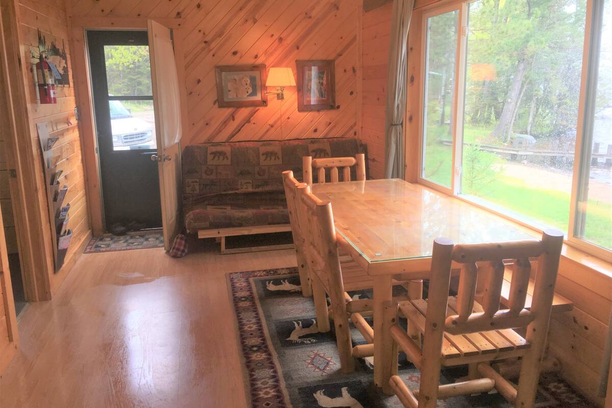 A wooden cabin interior with a dining table, chairs, a sofa, and a window overlooking a forest.