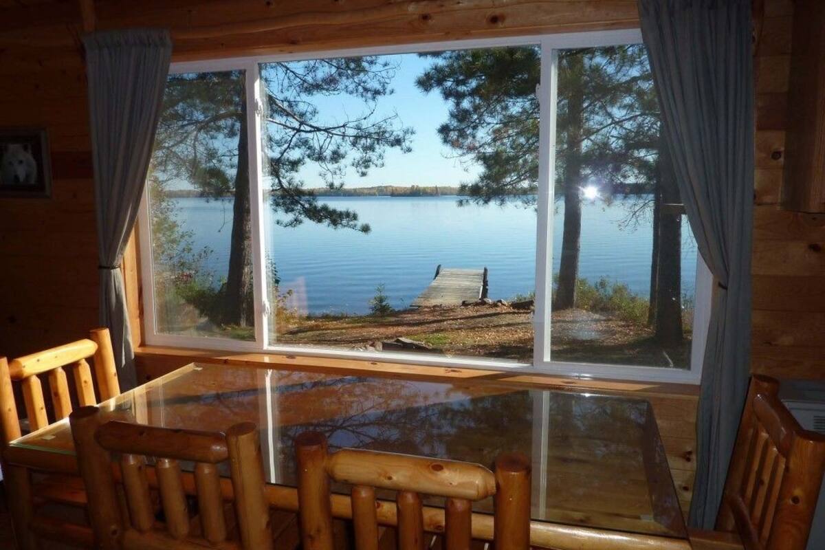 A wooden cabin with a glass dining table and chairs, a large window with curtains, and a view of a lake with a dock.